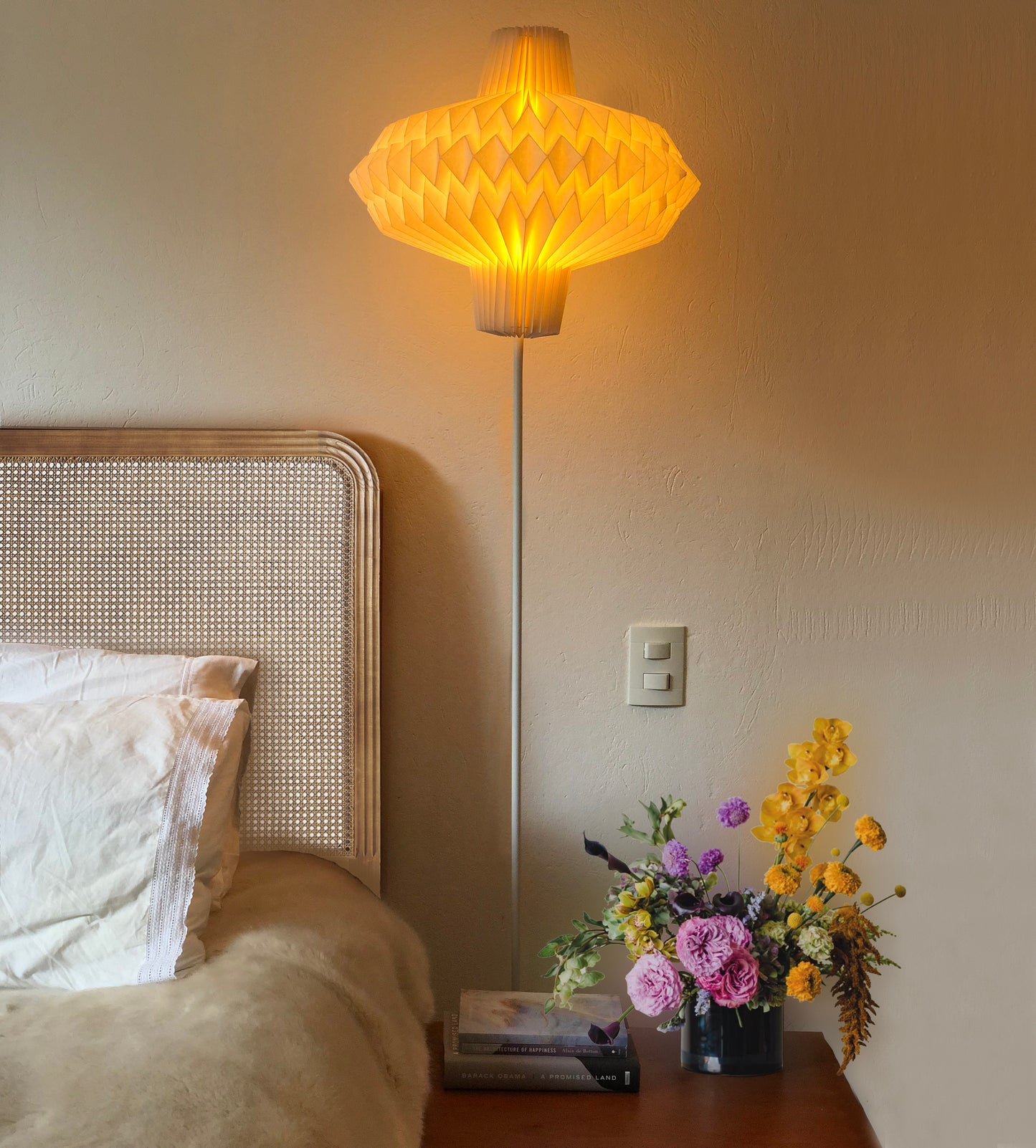 Plié Lights, lámpara de pie o lámpara de piso con base metálica blanca. Caperuza doblada a mano hecha en papel con técnica en origami. La lámpara está puesta en una habitación. A la izquierda se ve parte de una cama y abajo en el centro hay una mesa de noche con un florero. Diseño colombiano, lámpara de pie en Colombia. Venta disponible en todo colombia