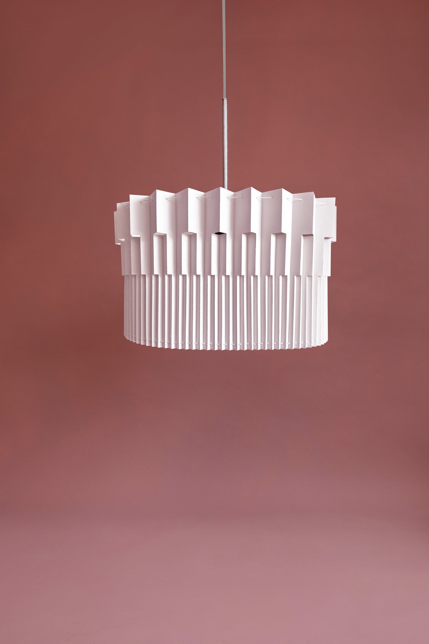 Lámpara colgante de Colombia. Plié Lights, referencia Museo de la colección monumentos. Fotografía de producto con fondo de color rosa. Caperuza escultórica estilo arquitectónico. El papel tiene cortes lo que permite genera movimiento entre luz directa e indirecta. Hecha en papel a mano. La base metálica es color blanco y cable transparente.