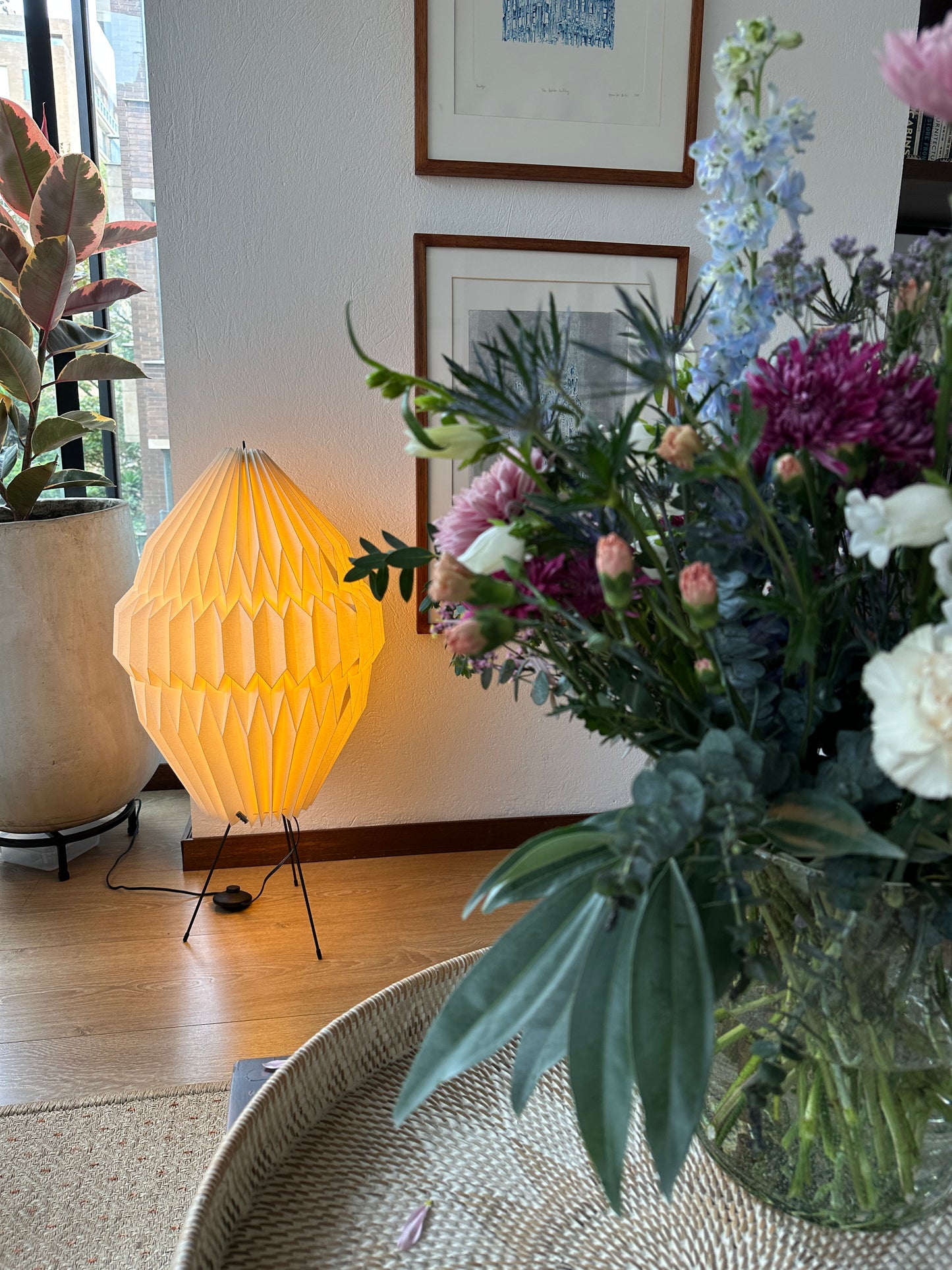 Plié Lights, lámpara de piso altura baja referencia Aries, hecha a mano, caperuza hecha en papel plegado a mano. Soporte metálica y cable transparente. Acabado blanco. Producto hecho en Colombia. Diseño Colombiano. igura geométrica muy interesante. Ideal para iluminar y darle calidez a los espacios. La lámpara está encendida. En el primer plano de la fotografía hay un florero y en el fondo a mano izquierda está la lámpara.