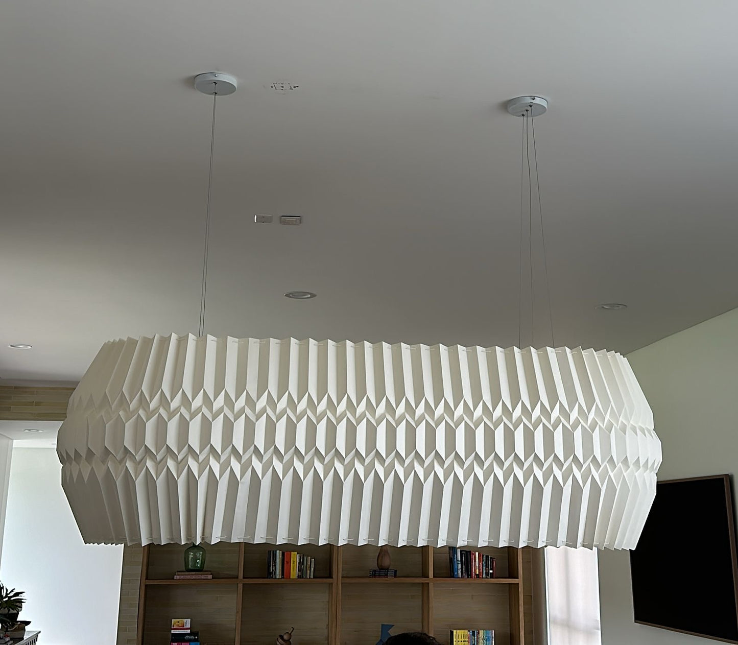 Lámpara colgante de Colombia. Plié Lights, referencia Tauro, un fondo de color rosa. Caperuza escultórica con diseño geométrico rectangular con ángulos redondeados. El diseño genera luz directa hacia arriba y hacia abajo. Hecha en papel a mano. La base metálica es color nblanco y cable guaya.