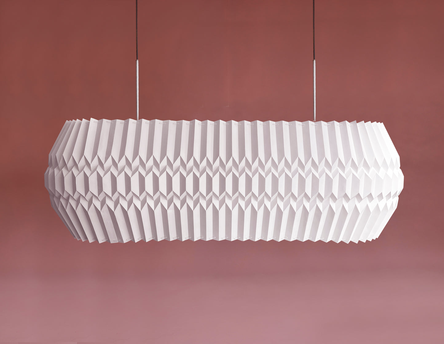 Lámpara colgante de Colombia. Plié Lights, referencia Tauro, un fondo de color rosa. Caperuza escultórica con diseño geométrico rectangular con ángulos redondeados. El diseño genera luz directa hacia arriba y hacia abajo. Hecha en papel a mano. La base metálica es color nblanco y cable guaya.