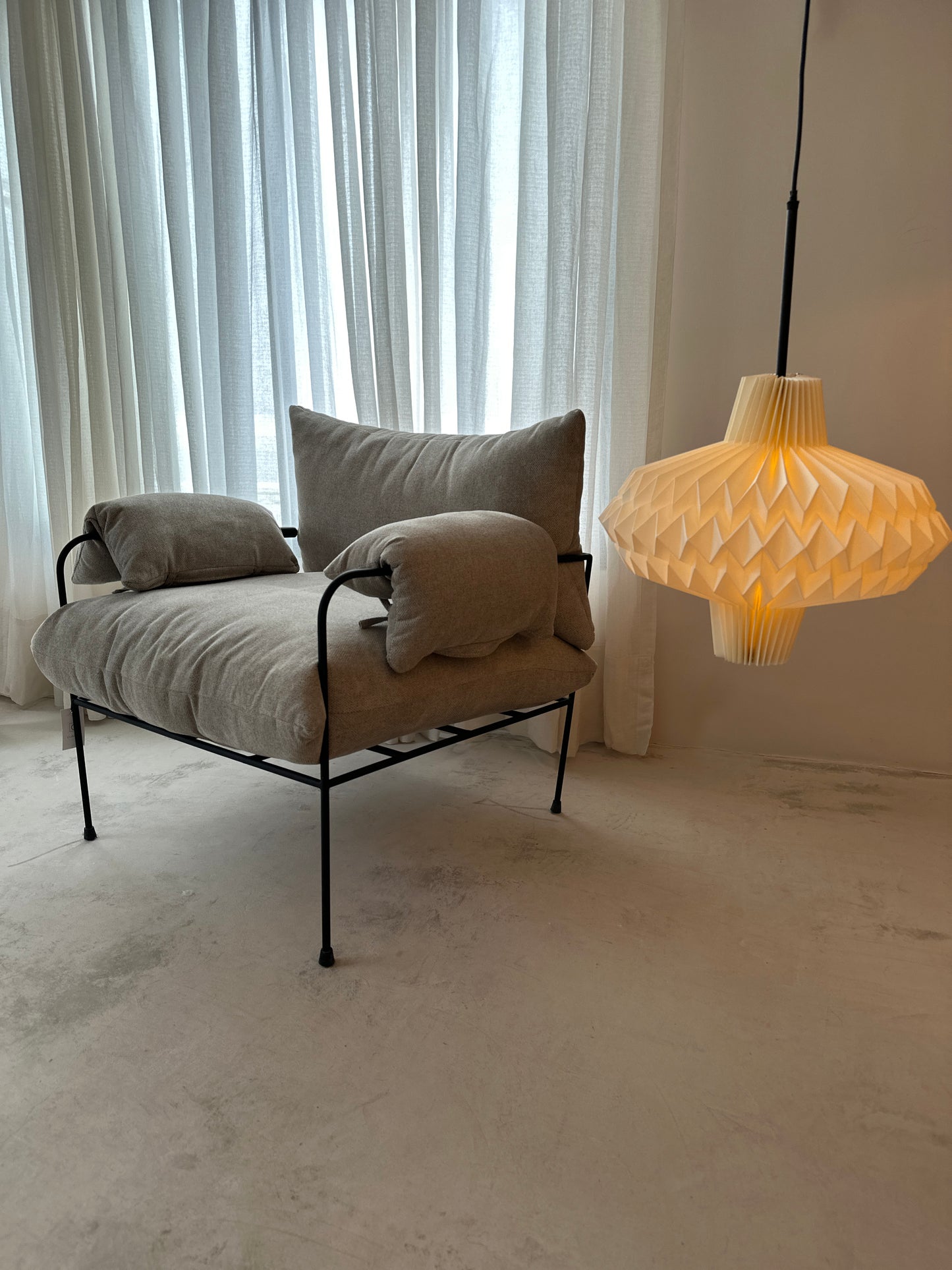 Plié Lights, lámparas hechas en Colombia. Caperuzas o pantalladas para iluminación hechas con papel doblado técnica de origami. La lámpara colgante referencia Antares está en un espacio interior, el showroom, al lado de una poltrona acolchonada y muy cómoda. Espacio cálido.