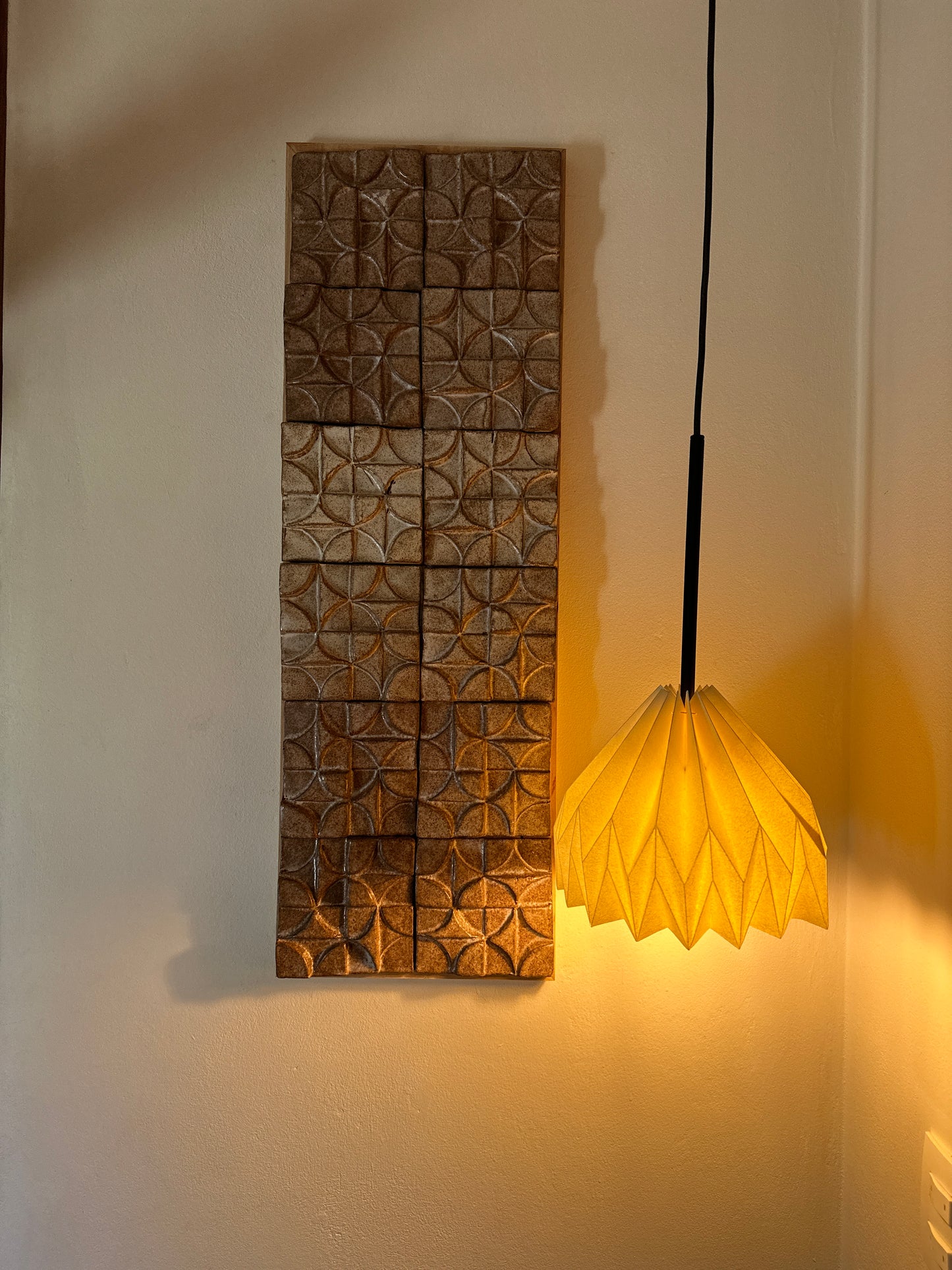 Lámpara colgante de Colombia. Plié Lights, referencia Gemini. Fotogragía de producto sobre un fondo de color rosa. Caperuza escultórica con diseño geométrico hecha en papel a mano. La lámpara hecha en Colombia está prendida. Al lado se ve un cuadro en relieve geométrico.