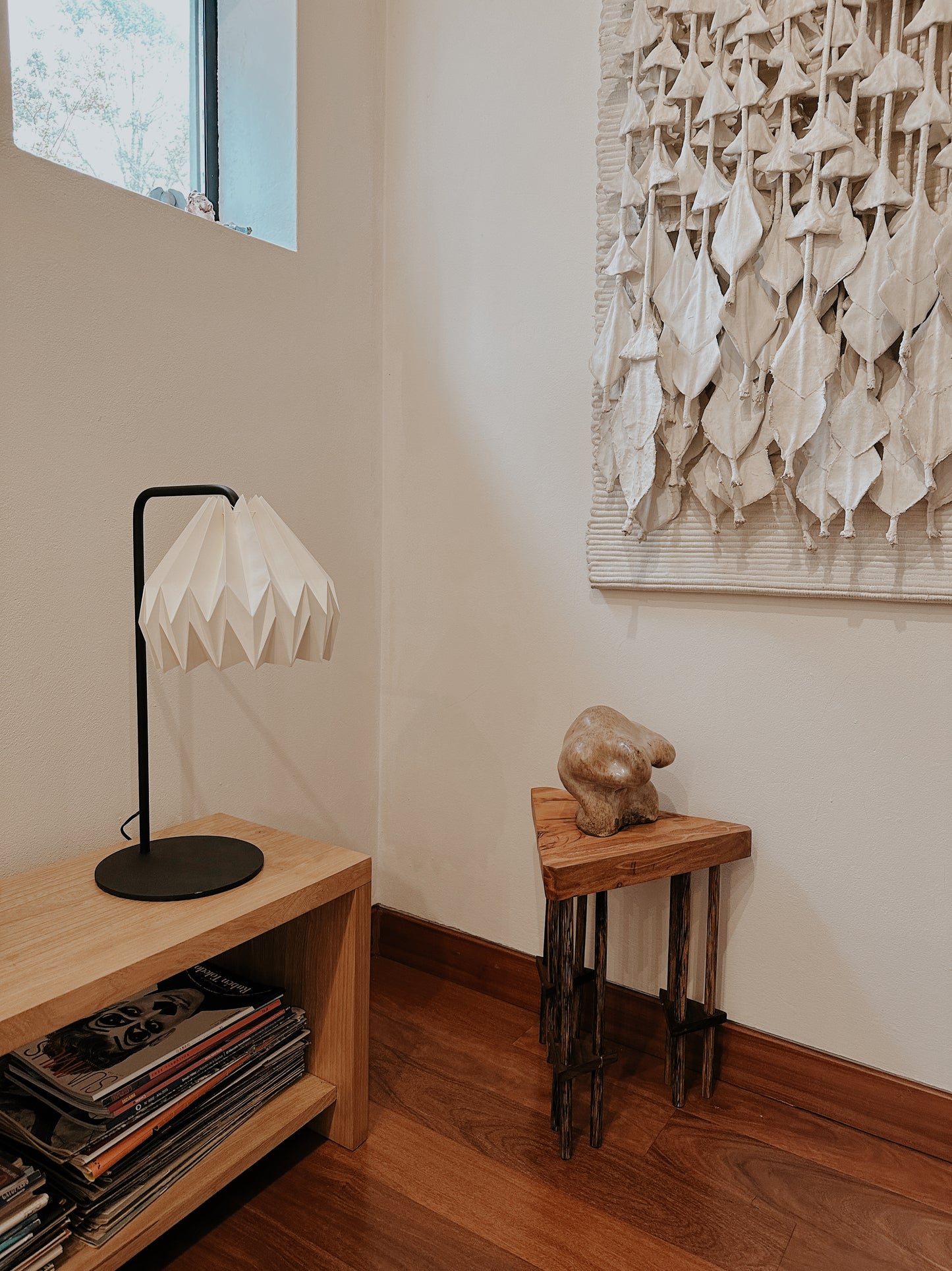 Plié Lights, lámpara de mesa hecha en Colombia, venta nacional. La lámpara está ubicada en un corredor, puesta sobre una banca con revistas, al lado hay otra mesita con una esctultura y un cuadro colgado en la parte superior derecha de la foto. La caperuza o pantalla de la lámpara está hecha a mano en papel plegado. Origami. Diseño Colombiano exclusivo de Plié Lights.