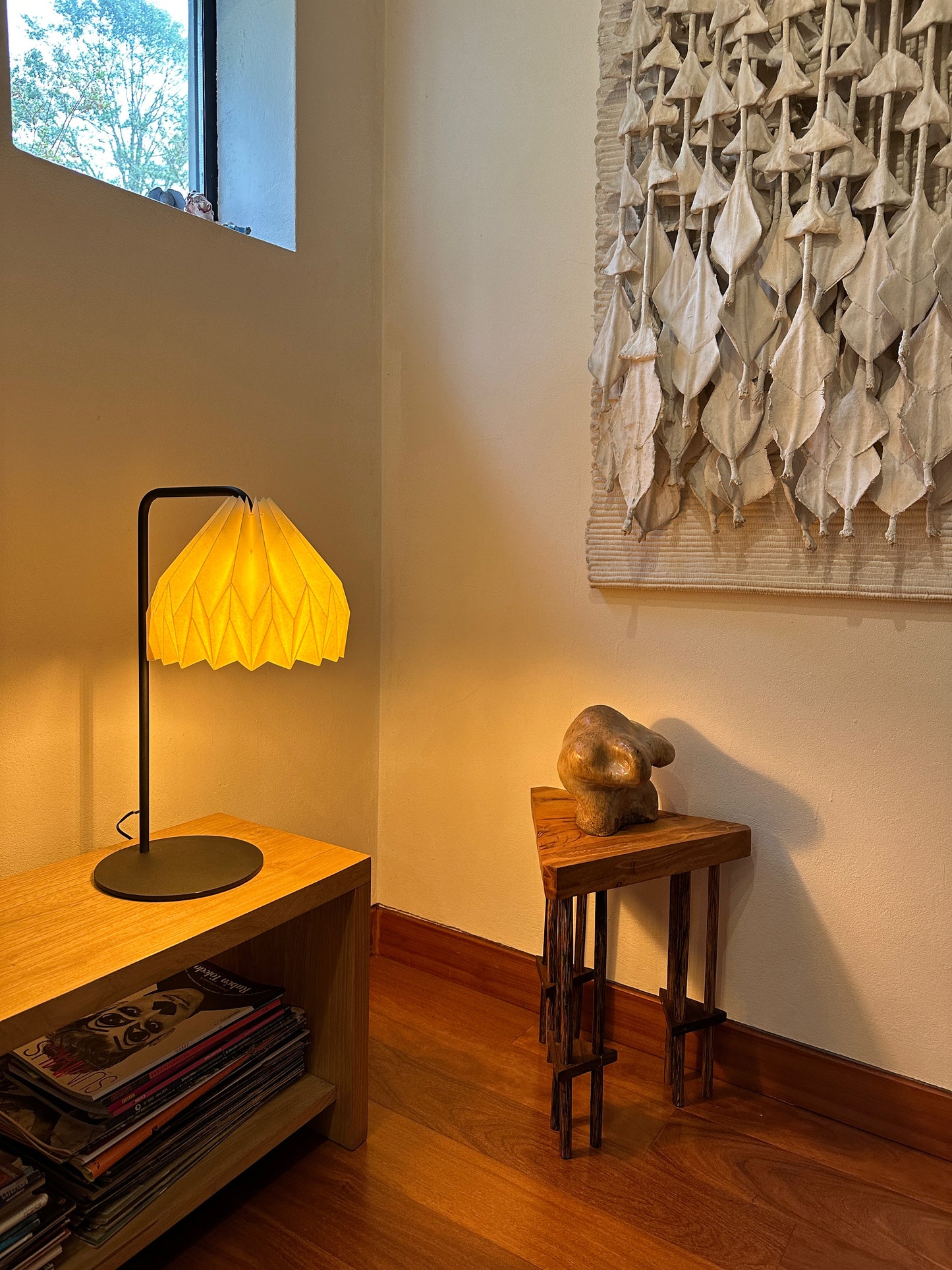 Plié Lights, lámpara de mesa hecha en Colombia, venta nacional. La lámpara está ubicada en un corredor, puesta sobre una banca con revistas, al lado hay otra mesita con una esctultura y un cuadro colgado en la parte superior derecha de la foto. La caperuza o pantalla de la lámpara está hecha a mano en papel plegado. Origami. Diseño Colombiano exclusivo de Plié Lights. Referencia Gemini. A diferencia de la fotografía anterior, en esta foto la lámpara está prendida.