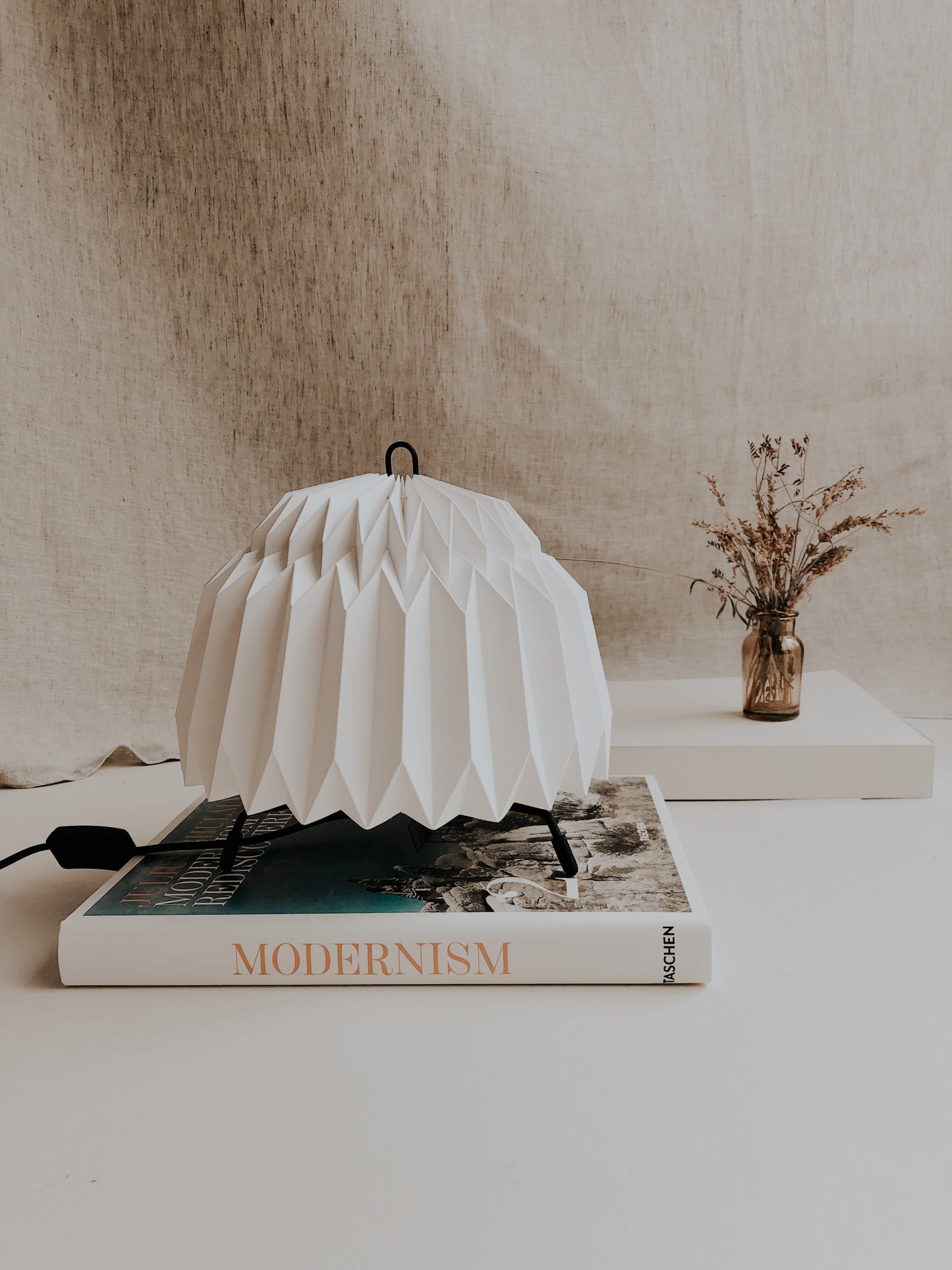 Lámpara de mesa marca Plié, referencia Leo, fabricada con base metálica y caperuza de papel plegado. Idea para espacios pequeños e iluminar los espacios con un ambiente especial. Diseño colombiano, hecha a mano en Colombia. Caperuca hecha en origami, doablada a mano con papel. La lámpara está puesto encima de un libro y hay un pequeño florero detrás.