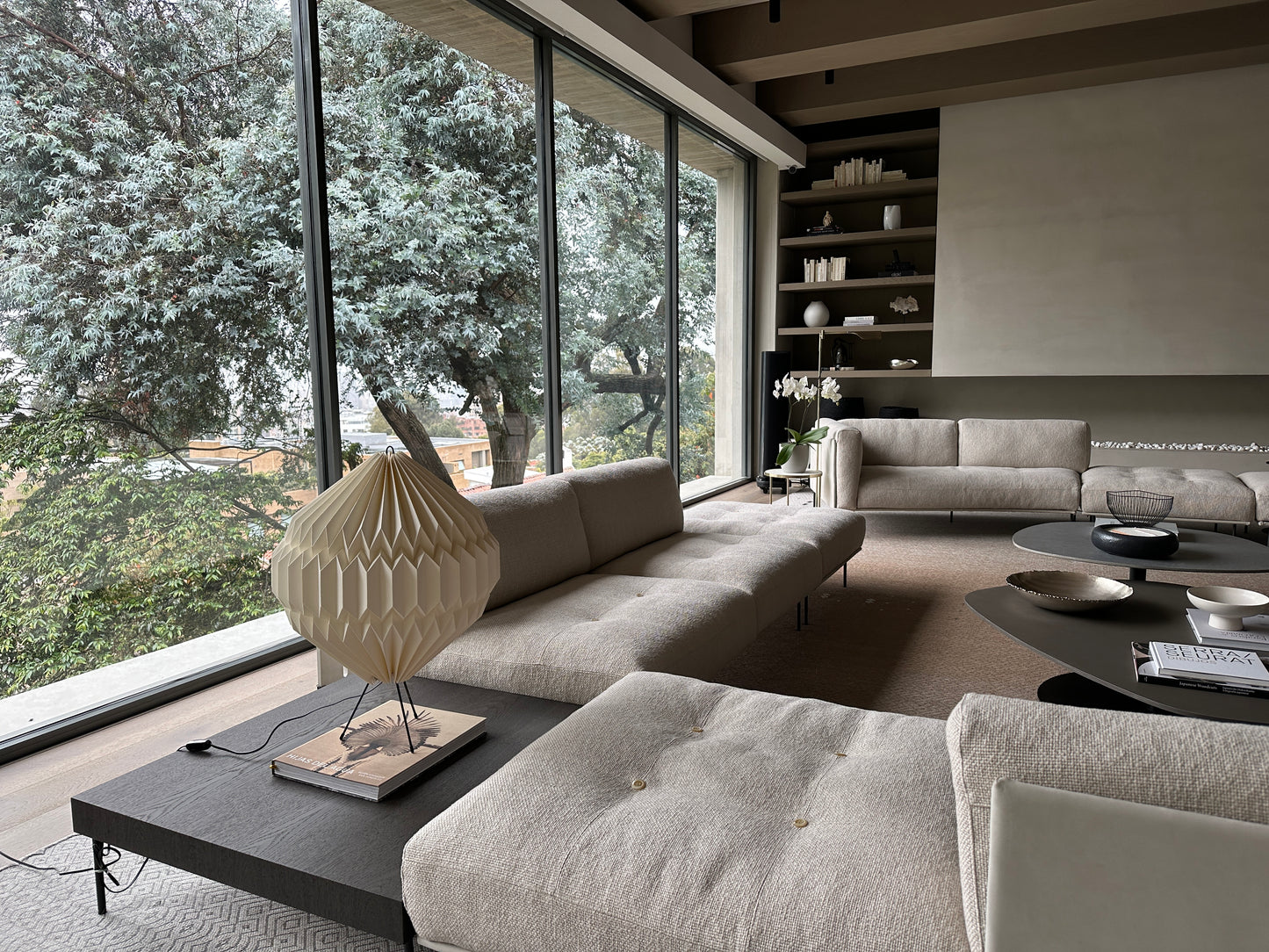 Lámpara Plié Lights, lámpara de mesa o piso bajita referencia Luna. La caperuza es hecha con papel doblado como origami. La lámpara está puesta en una mesa auxiliar al lado de unos sofás. Puesta en la sala o salón social de una casa en Bogotá.