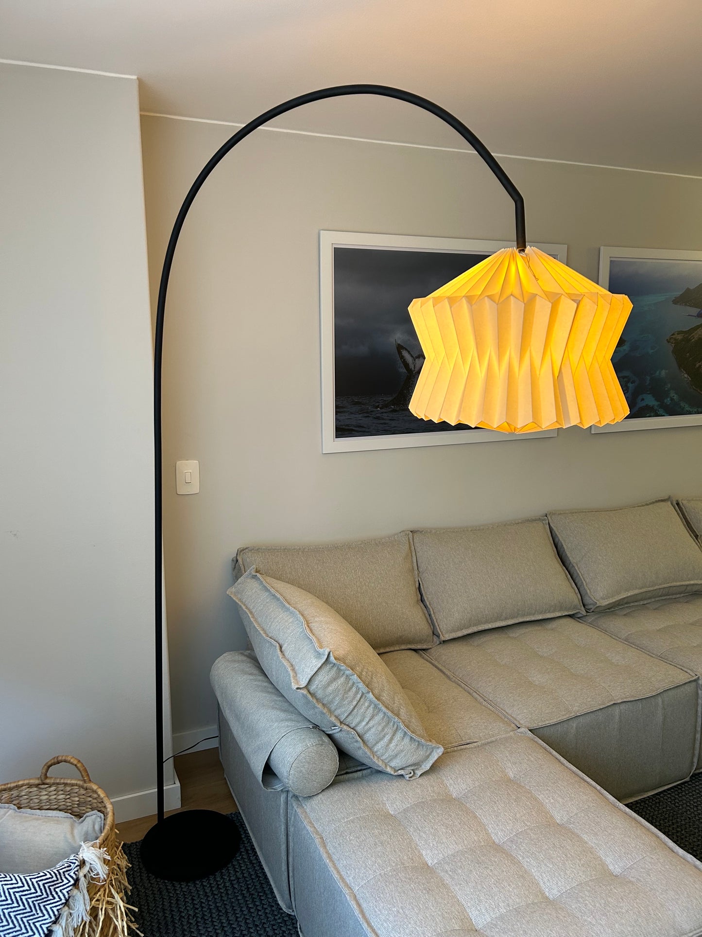Plié Lights, lámpara de pie o lámpara de piso hecha a mano en Colombia. Referencia Altair. La lámpara está puesta en un estudio o family room. Se ve un sofá en L y una canasta con cobijas. La caperuza o pantalla de la lámpara está hecha en origami de papel doblado a mano por artesanos Colombianos.