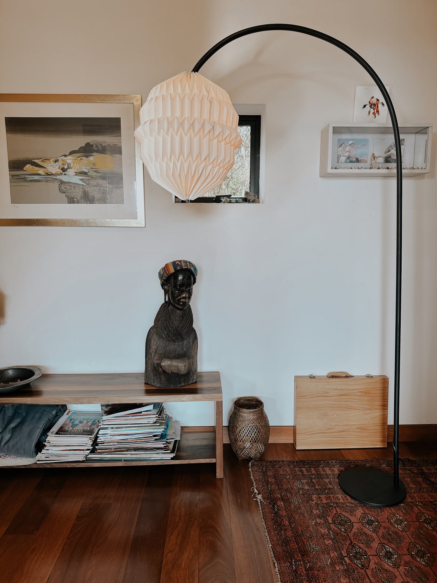 Lámpara de piso Plié Lights. Esta lámparas hechas en Colombia es una fotorafía de producto. La lámpara tiene una base metálica color negro y tiene una forma en arco, lo que permite crear el efecto de una lámpara colgante pero con una base de piso. La lámpara está puesta en un espacio interior, hay una banca con una escultura y unas revistas abajo. El piso es de madera.