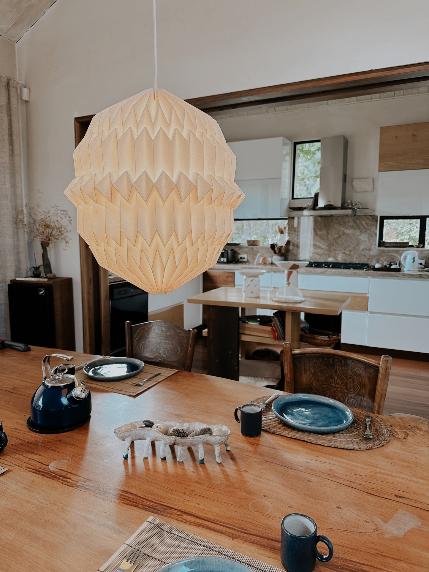 Plié Lights, lámpara colgante referencia Aries, hecha a mano, caperuza hecha en papel plegado a mano. Soporte metálica y cable negro. Acabado negro. Producto hecho en Colombia. Diseño Colombiano. igura geométrica muy interesante. Ideal para iluminar y darle calidez a los espacios. La lámpara está puesta encima de una mesa de comedor detrás se ve la cocina.