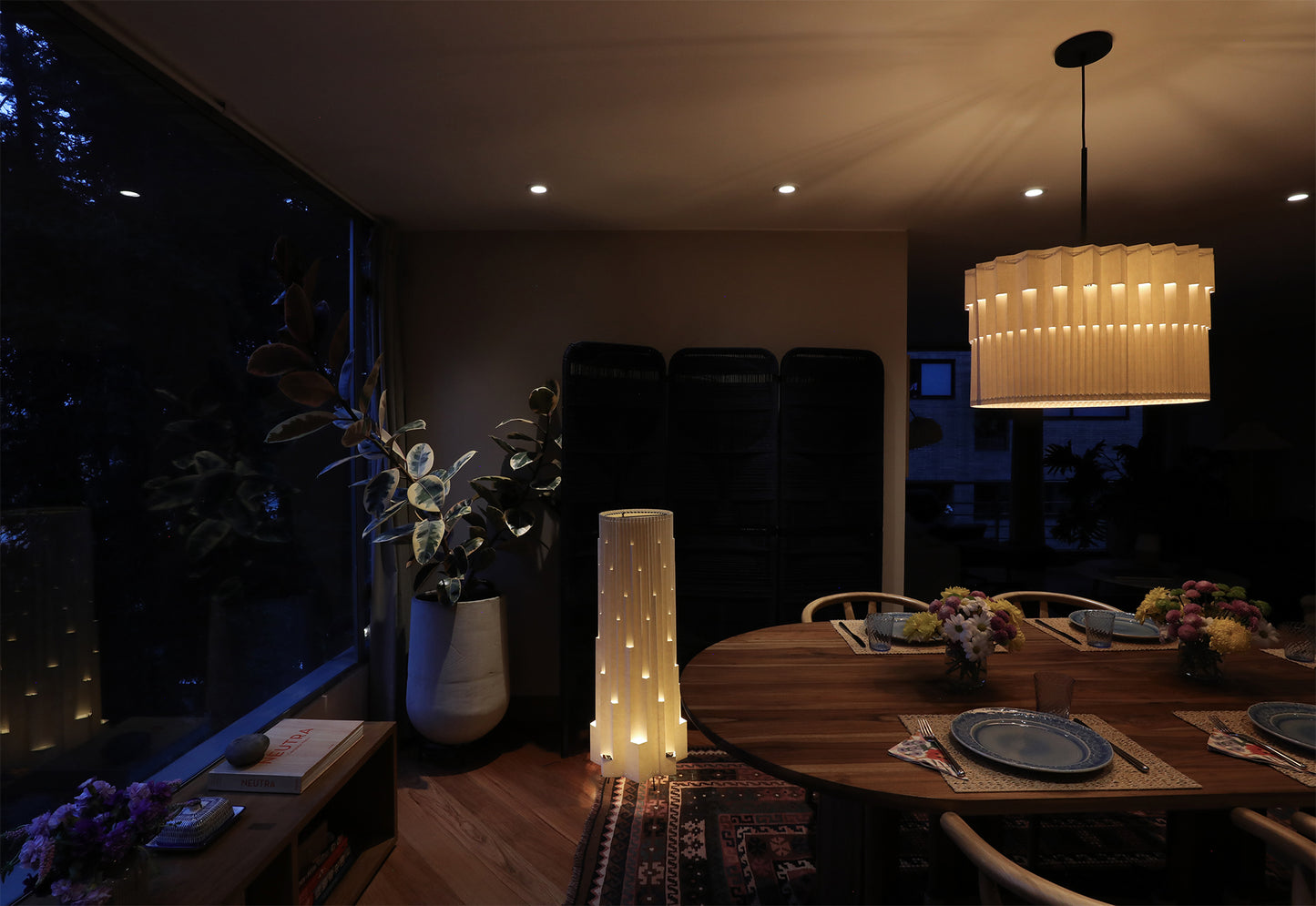 Lámpara de piso de Colombia. Plié Lights, referencia Torre de la colección monumentos. Fotografía en sitio ubicada e un comedor, en un primer plano podemos ver otra lámpara de Plié Lights de la colección monumentos referencia Museo. Caperuza escultórica estilo arquitectónico. El papel tiene cortes lo que permite genera movimiento entre luz directa e indirecta. Hecha en papel a mano. La base metálica es color negro y cable negro. La foto fue tomada al final del día.