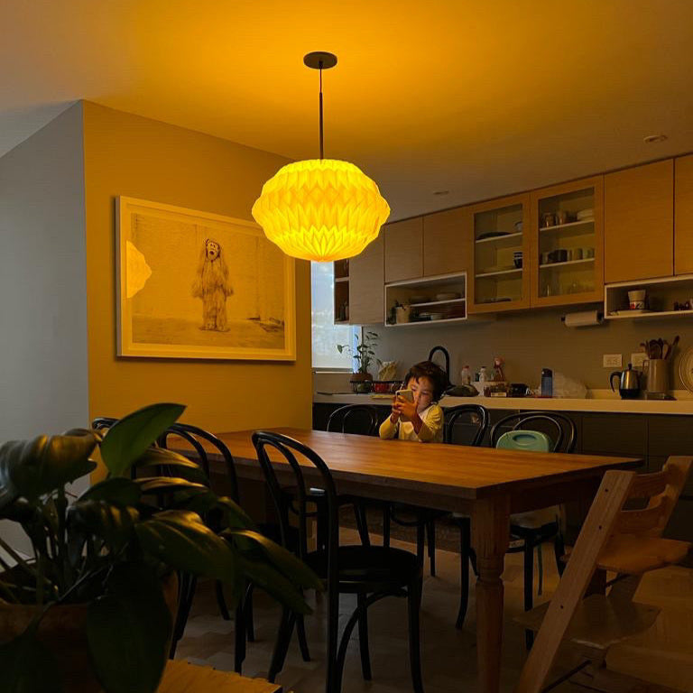 Plié Lights_Lámpara de techo referencia Libra, la caperuza es hecha con papel en origami, doblada y hecha a mano. Diseño de lámparas de Colombia. La lámpara colgante está colgada encima de una mesa de comedor, en la mesa hay un niño. Atrás se ve la cocina.
