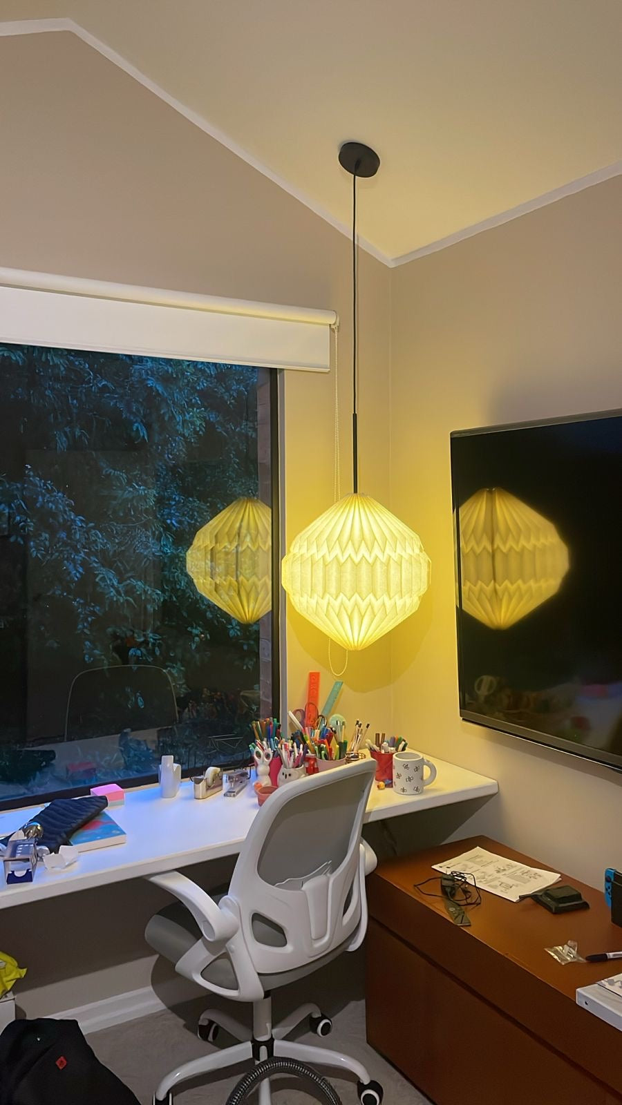 Plié Lights, lámpara colgante referencia luna, hecha a mano, caperuza hecha en papel plegado a mano. Soporte metálica y cable transparente. Acabado blanco. Producto hecho en Colombia. Diseño Colombiano. Figura geométrica muy interesante. Ideal para iluminar y darle calidez a los espacios. Instalada en un estudio, donde hay un escritorio, una silla de trabajo y una ventana en el fondo con un árbol.