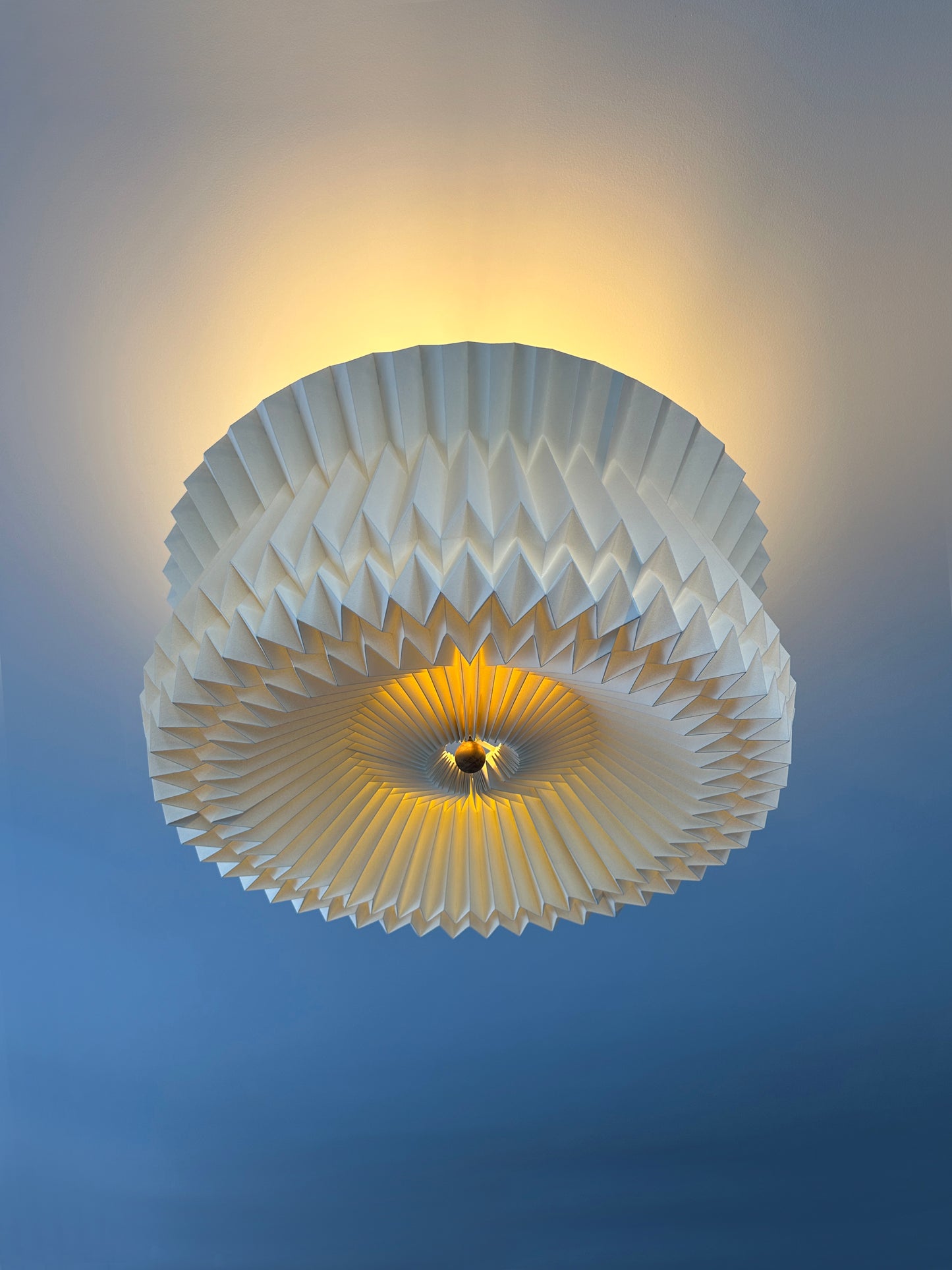 Lámpara colgante de Colombia. Plié Lights, referencia Pegaso fotografía de producto en fondo de color rosa. Caperuza escultórica con diseño geométrico. La parte superior de la lámpara es abierta, lo que genera luz directa hacia el techo. La parte inferior es cerrada y está un poco metida con respecto al borde de la caperuza. Hecha en papel a mano. La base metálica es color negro y cable negro. Vista tomada de abajo hacia arriba, se ven los detalles de la lámpara de la parte inferior de la caperuza.