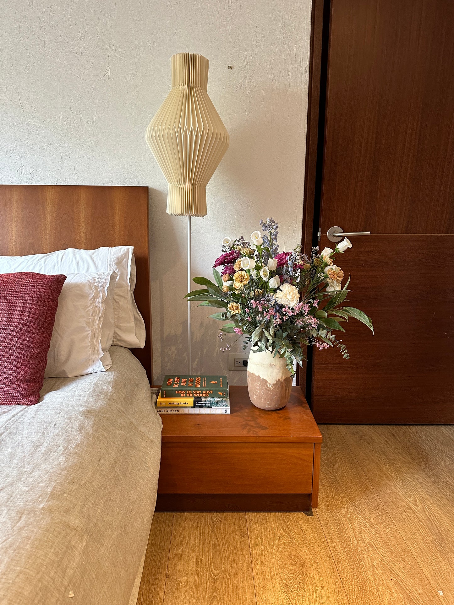 Plié Lights_lámpara de piso referencia Hydra. Lámpara de 155 cm de alto, con caperiza en papel una figura geometrica hecha en orginami con base metálica negra. La lámpara está puesta al lado de una cama, enfrente de la lámpara hay una mesa de noche con libros y un florero. La lámpara está apagada. A mano ederecha se ve una puerta. .