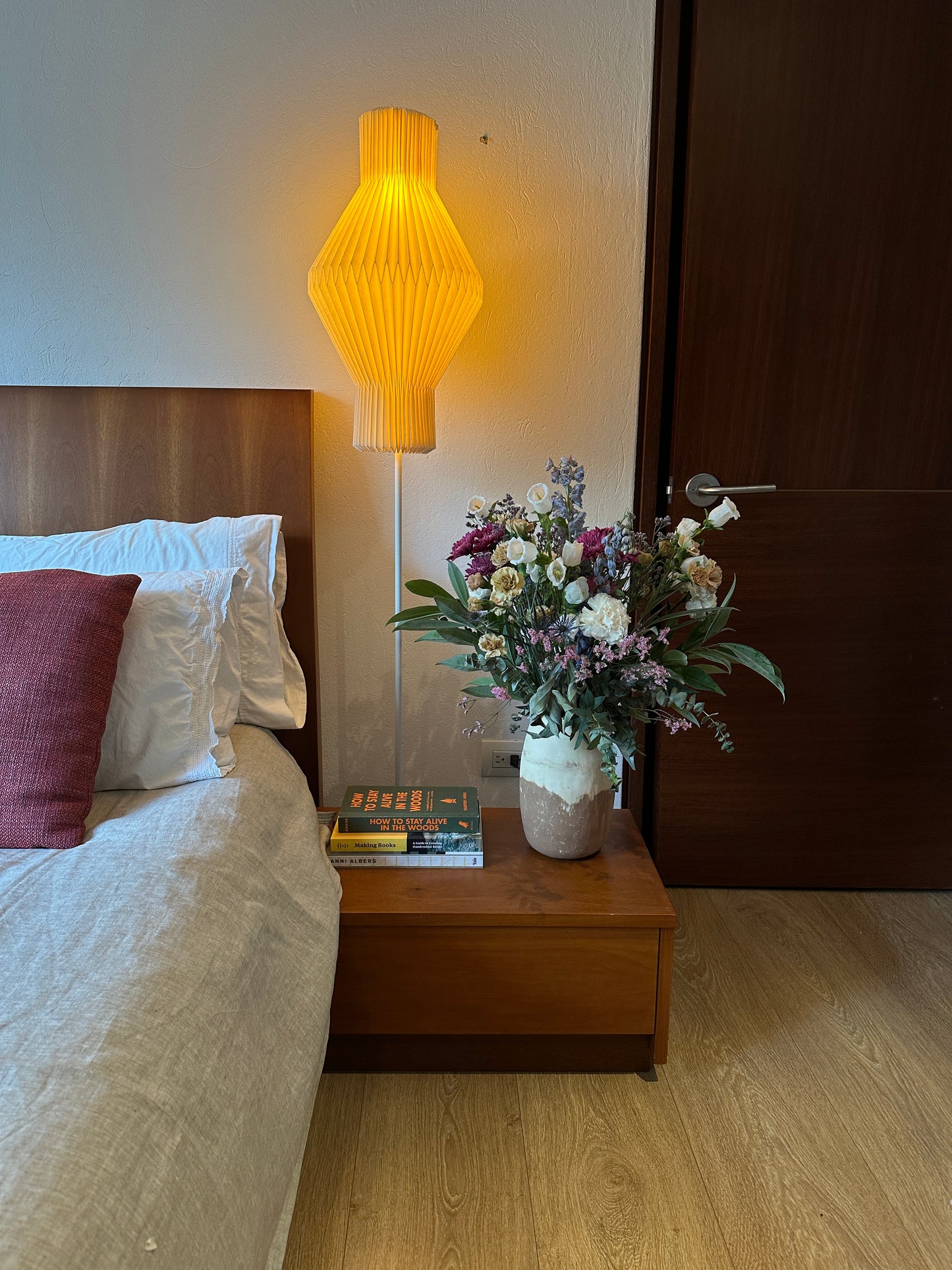 Plié Lights_lámpara de piso referencia Hydra. Lámpara de 155 cm de alto, con caperiza en papel una figura geometrica hecha en orginami con base metálica negra. La lámpara está puesta al lado de una cama, enfrente de la lámpara hay una mesa de noche con libros y un florero. La lámpara está prendida y genera una iluminación cálida, una lámpara estilo japonés.