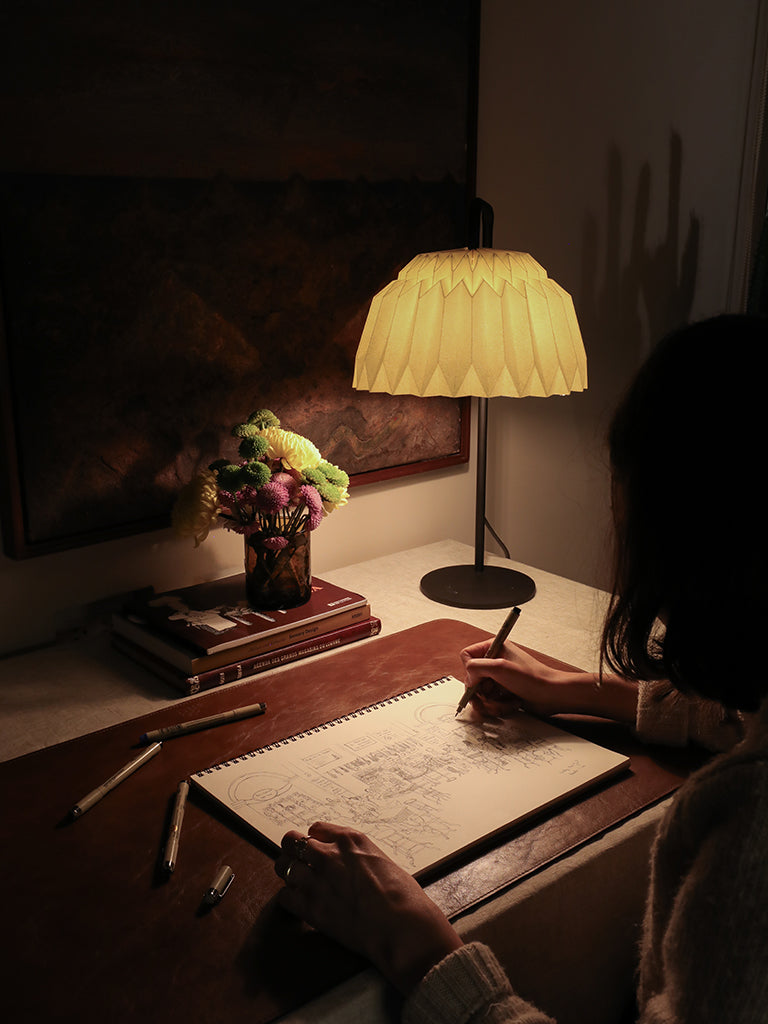 Lámpara Lyra de mesa fabricada con base metálica y caperuza hecha en papel plegado. Perfecta para iluminar espacios pequeños y dar un brillo a los espacios. Lámparas hechas en colombia. Fotografía de la lámpara puesta sobre un escritorio, donde se ve la lámpara iluminando el cuadernos de una persona que dibuja a mano con esfero y papel. Al fondo hay unos libros y un florero.