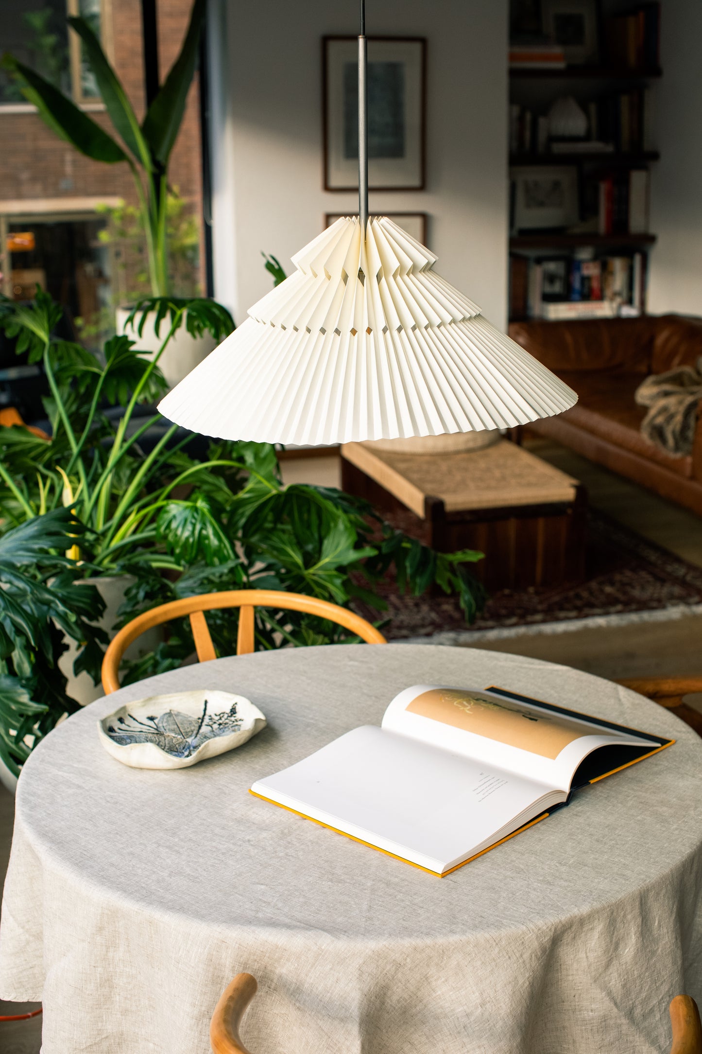 Lámpara marca Plié, referencia Andrómeda caperuza fabricada con papel plegado y base metálica con un diseño colombiano y figura geométrica muy interesante. Ideal para iluminar y darle calidez a los espacios. En la escena está ubicada debajo de una mesa de comedor.