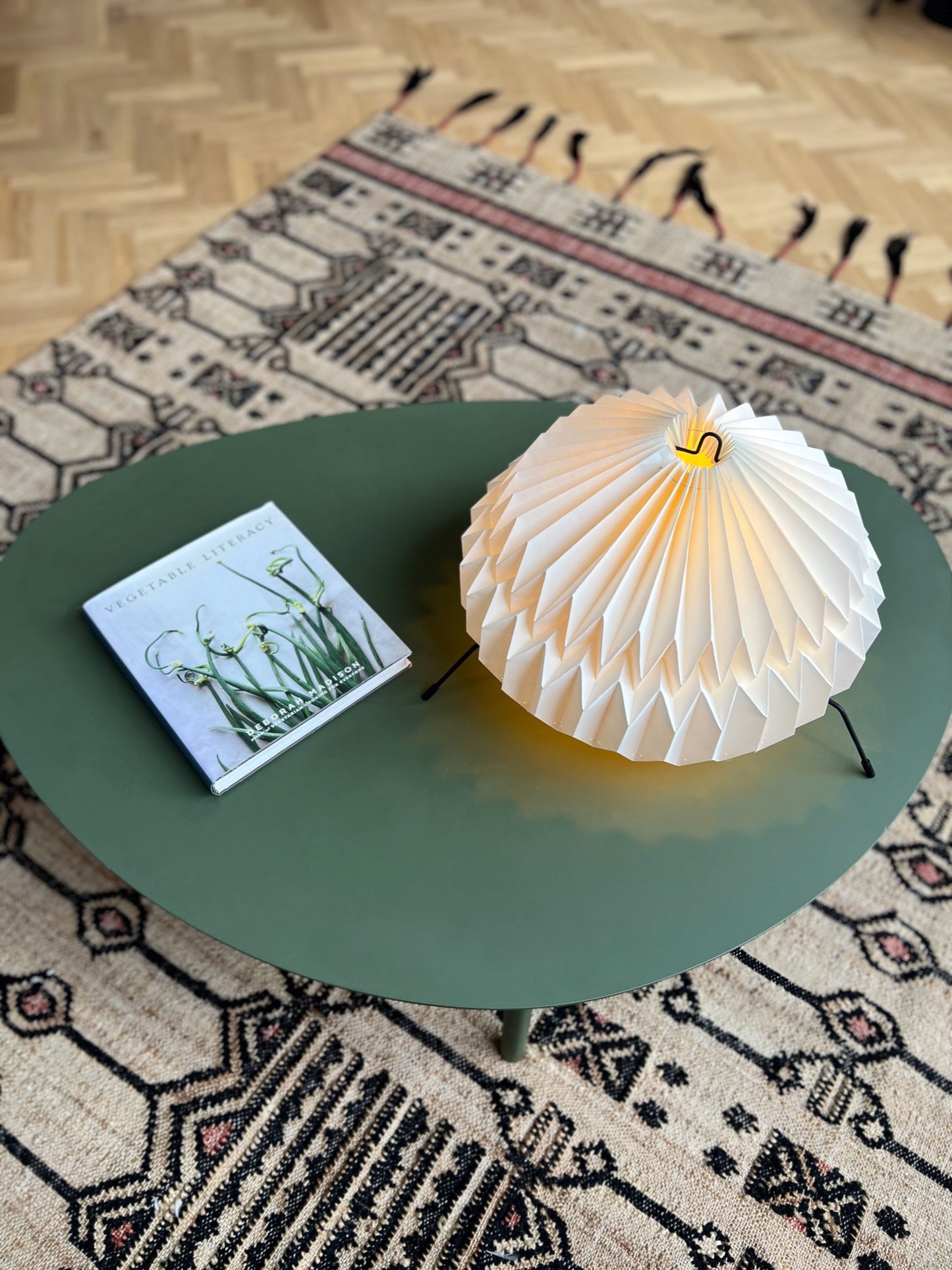 Lámpara Plié de mesa o lámpara de piso referencia Atria, fabricada con base metálica y caperuza hecha con papel plegado. Diseño colombiano. La lámpara está puesta sobre una mesa, se puede comparar el tamaño con un libro y en el piso se visualiza un tapete.