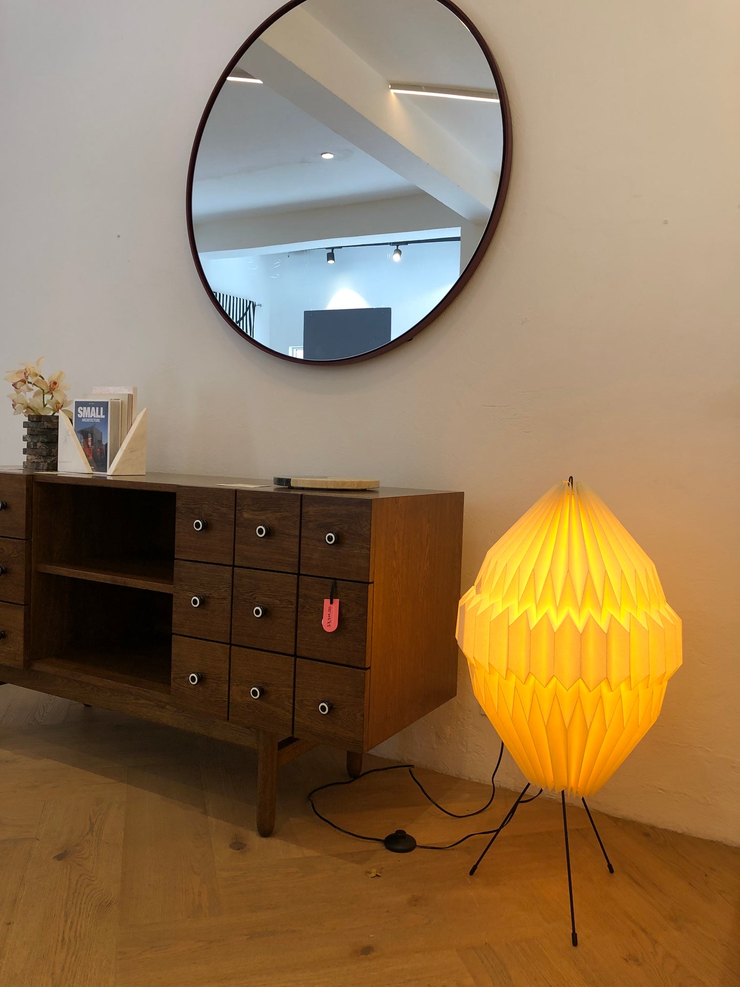 Plié Lights, lámpara de piso altura baja referencia Aries, hecha a mano, caperuza hecha en papel plegado a mano. Soporte metálica y cable transparente. Acabado blanco. Producto hecho en Colombia. Diseño Colombiano. igura geométrica muy interesante. Ideal para iluminar y darle calidez a los espacios. La lámpara está encendida y está al lado de un consola con un espejo.
