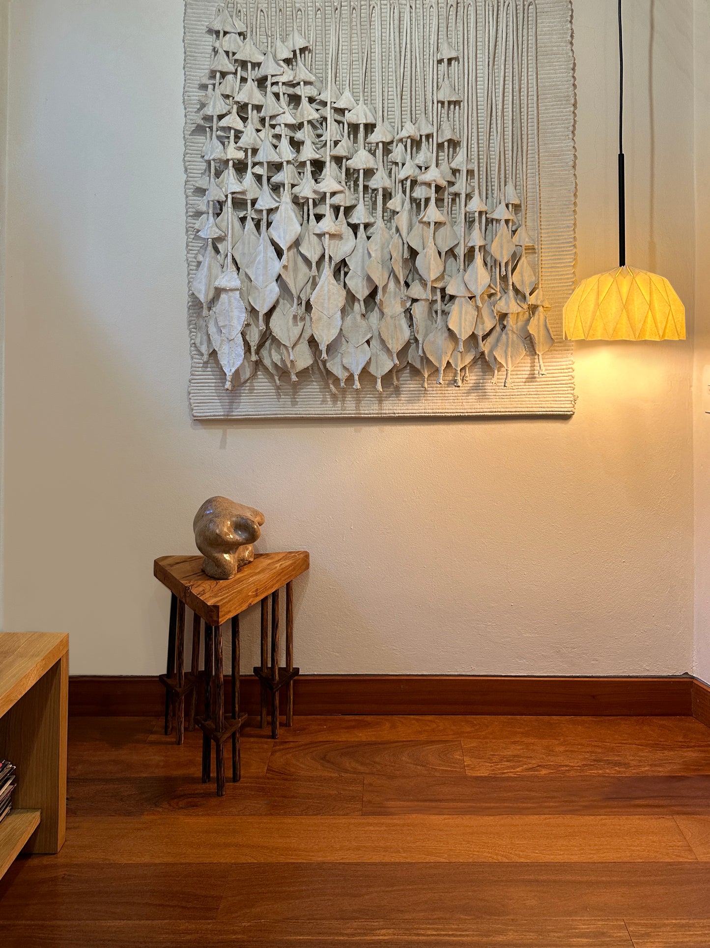 Lámpara colgante hecha en Colombia, referencia Fenix. Fotografía de producto puesta en un espacio. La lámpara está iluminada y se puede ver la luz cálida que produce. Sobre la pared hay un cuadro escultórico, una mesita con otra escultura. La lámpara es hecha en papel. Plié Lights
