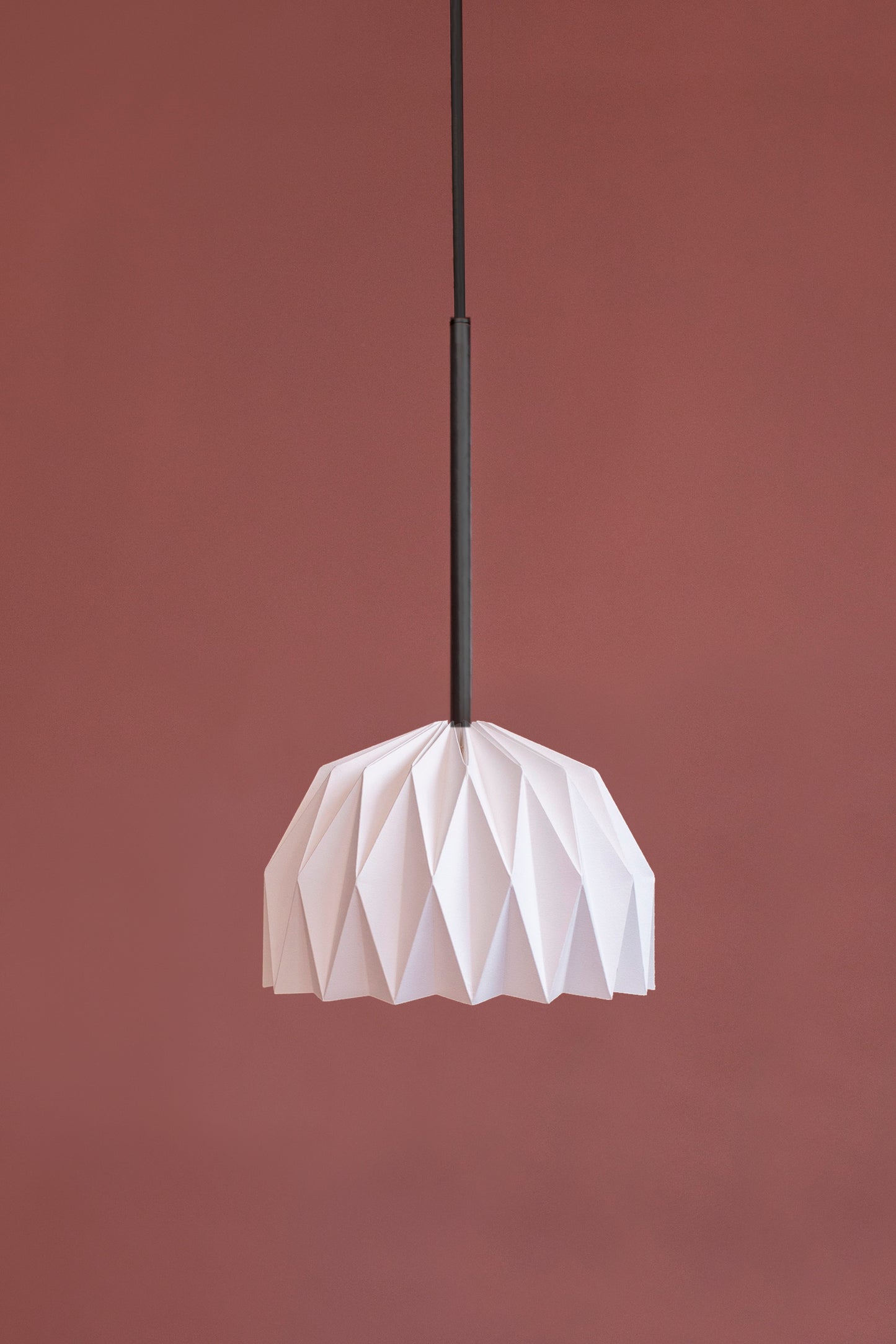 Lámpara colgante hecha en Colombia, referencia Fenix. Fotografía de producto, imagen de la lámpara sobre un fondo de color. Tiene una estructura colgante con un tubo metálico y cable color negro. La caperuza tiene una figura geometrica y la caperuza está hecha a mano en papel. Plié Lights