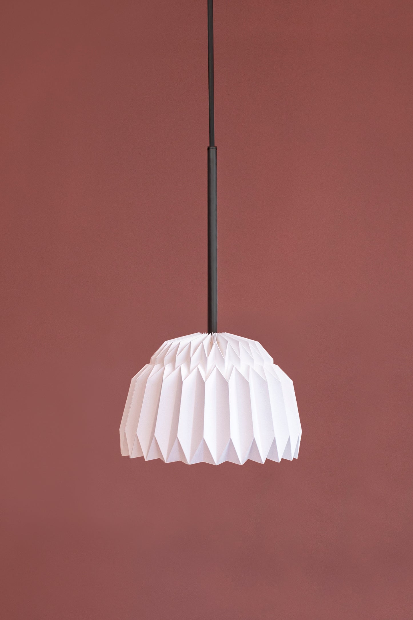 Lámpara colgante de Colombia. Plié Lights, referencia Leo, un fondo de color rosa. Caperuza escultórica con diseño geométrico en forma de semicirculo. Hecha en papel a mano. La base metálica es color negro y cable negro.