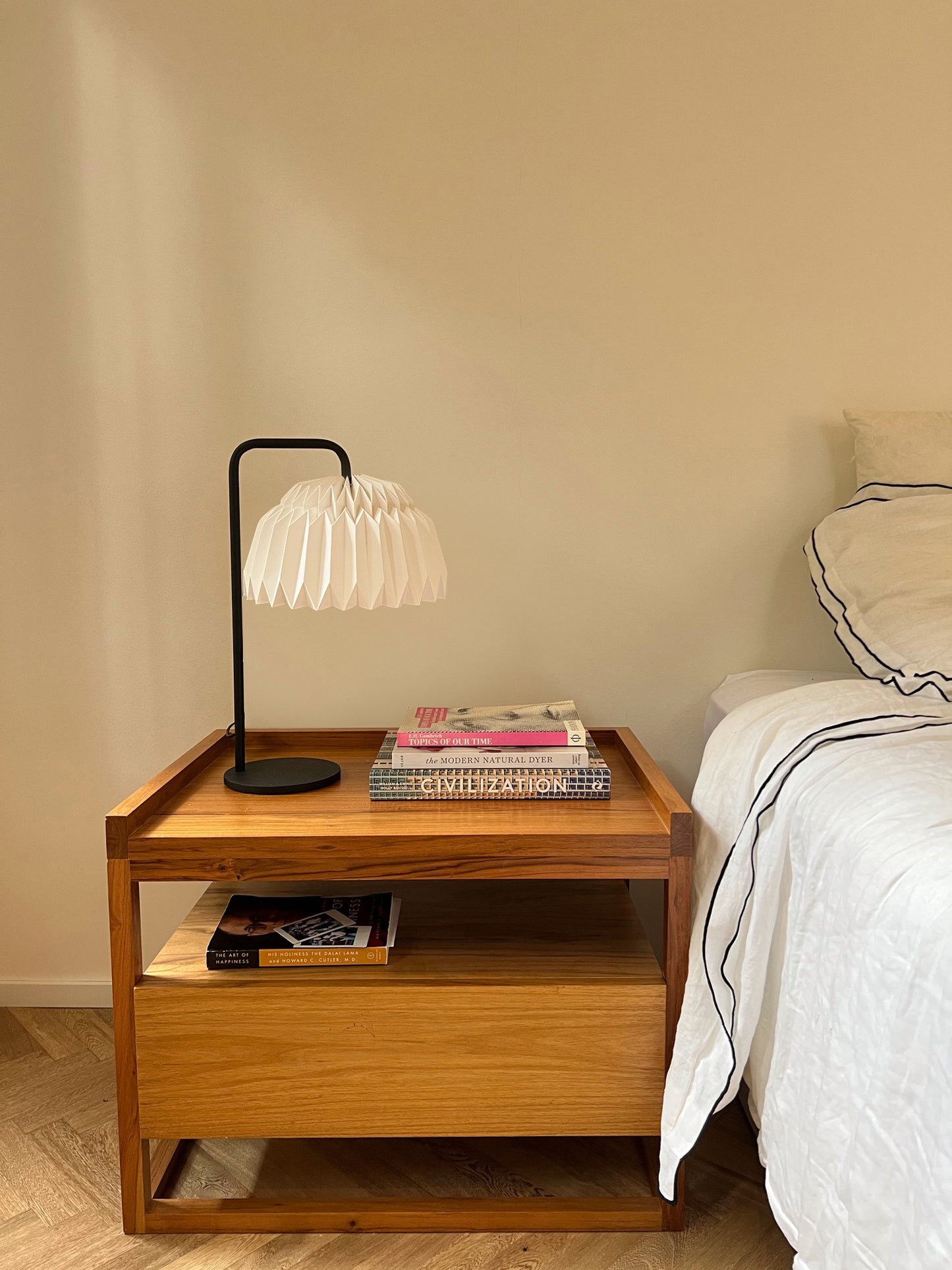 Lámpara de mesa Plié Lights. La lámpara está apagada y se visualiza cómo al no estar iluminada es un objeto funcional y escultórico. Caperuza hecha a mano de papel, con una figura geomtría semicirculo. Base metálica negra. La lámpara está puesta encima de una mesa de noche, en la mesa de noche hay libro y a la derecha está el borde una cama con un tendido en lino. La lámpara es diseñada y hecha a mano en Colombia. Lámpara de Colombia.