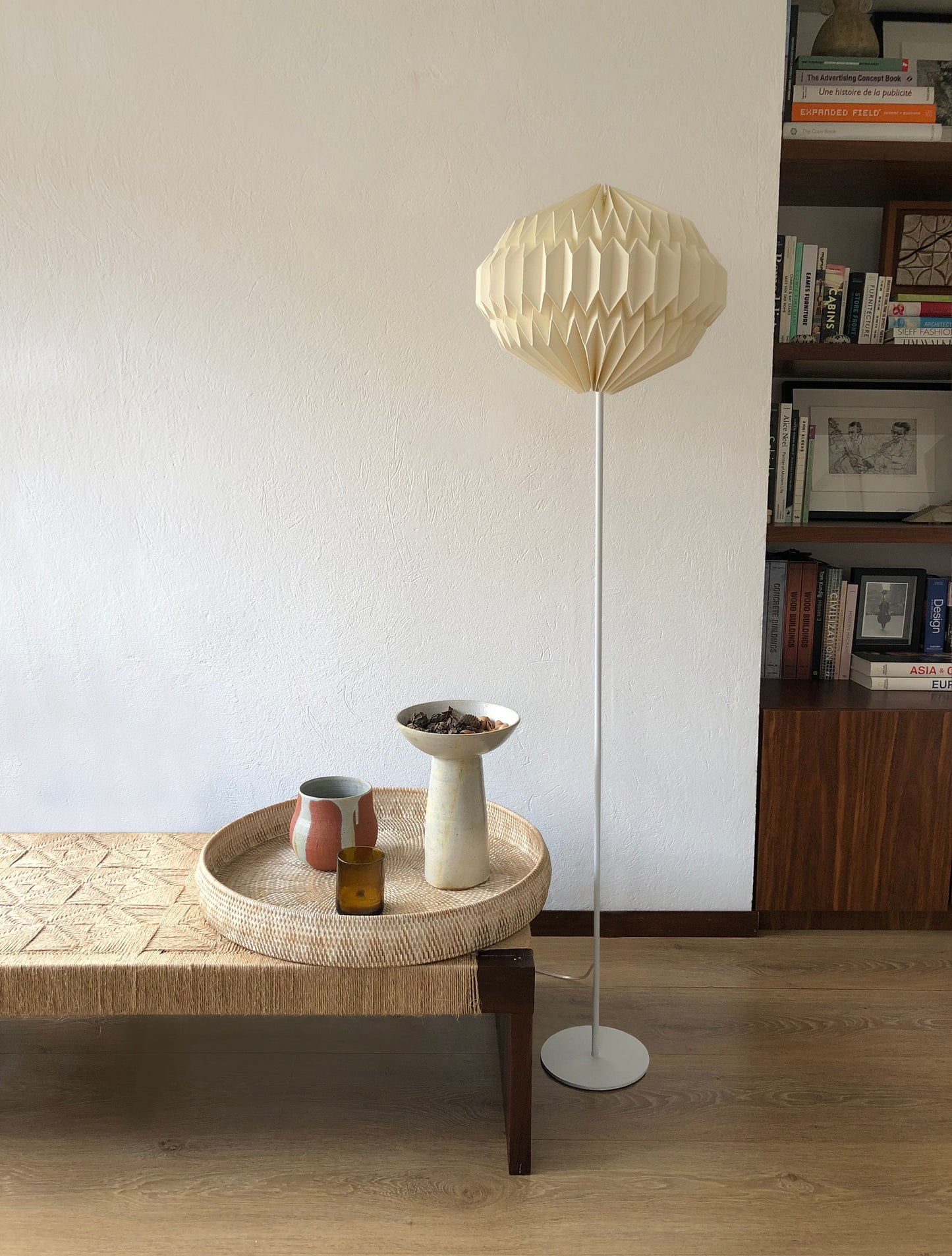 Plié Lights lámpara de piso referencia orión. Base metálica color blanco, caperuza simetríca redonda con un diseño geométrico. La lámpara está al lado de una mesa, que tiene un bandeja y unas cerámicas. Atrás a la derecha hay una biblioteca.
