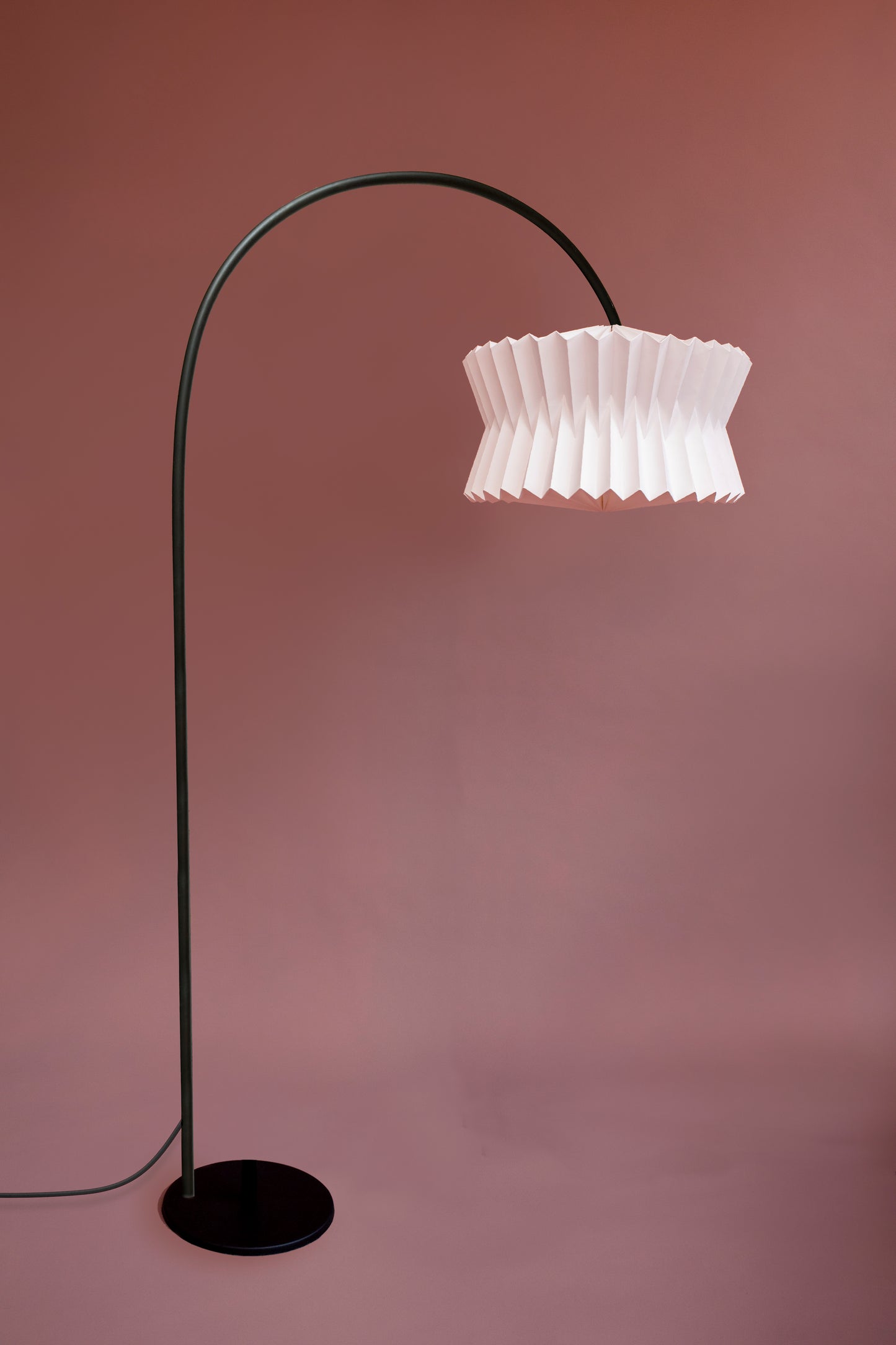 Plié Lights, lámpara de piso hecha en colombia. La base metálica es grande, tiene un arco lo que recrea un efecto de lámpara colgante. La caperuza es referencia Altair, tiene una forma de tambor con un diseño de pliegues en papel geométricos. Produce una iluminación cálida.