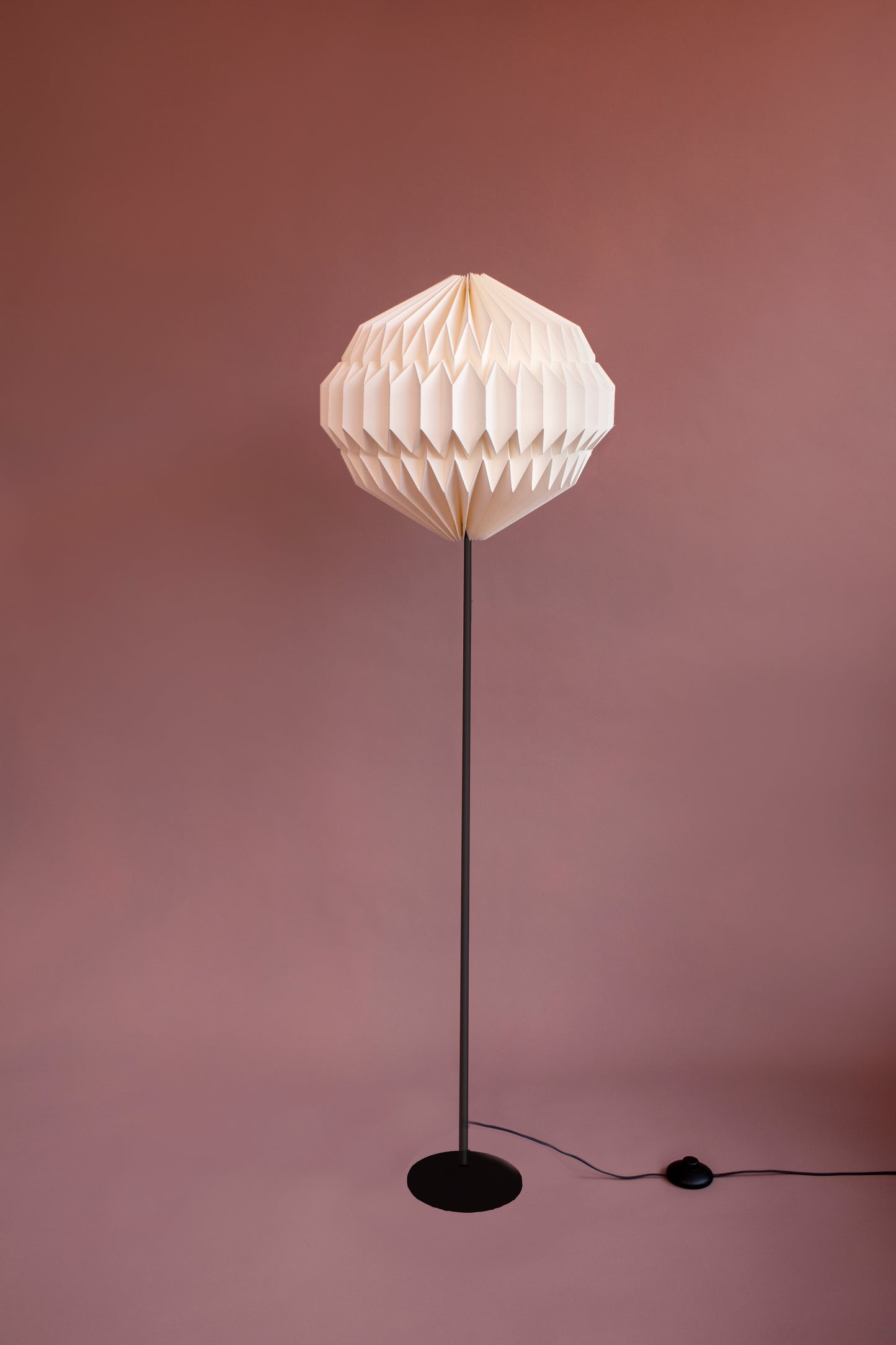 Plié Lights, lámpara de piso referencia Orión con base metálica color negro. Fotografía de producto sobre un fondo de color. La caperuza de papel con una geometría redonda es hecha a mano en Colombia. Lámparas para el hogar.