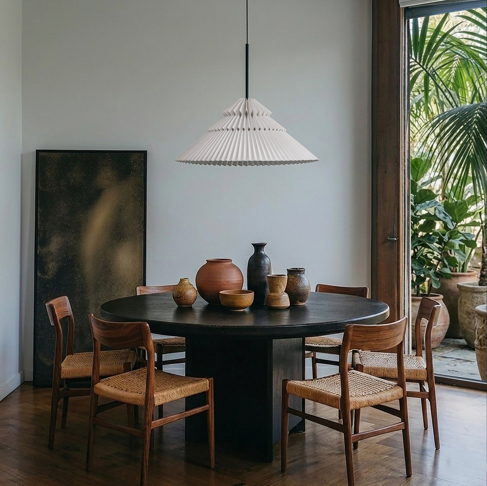 Lámpara marca Plié, referencia Andrómeda caperuza fabricada con papel plegado y base metálica con un diseño colombiano y figura geométrica muy interesante. Ideal para iluminar y darle calidez a los espacios. Fotografía en un comedor, la lámpara está colgando encima de la mesa, sobre la mesa hay unos jarrones en cerámica y hacia afuera se ve un balcón con matas. La mesa es de 6 persona y la escena es de día, la lámpara está apagada.