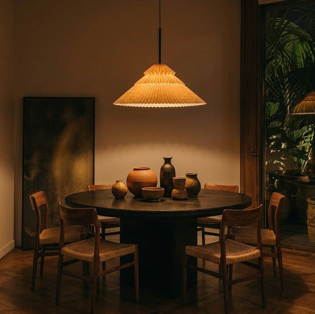 Lámpara marca Plié, referencia Andrómeda caperuza fabricada con papel plegado y base metálica con un diseño colombiano y figura geométrica muy interesante. Ideal para iluminar y darle calidez a los espacios. Fotografía en un comedor, la lámpara está colgando encima de la mesa, sobre la mesa hay unos jarrones en cerámica y hacia afuera se ve un balcón con matas. La mesa es de 6 persona y la escena es nocturna, la lámpara está prendida y el ambiente es cálido. 