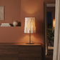 Fotografía de la lámpara Plié Lights, lámpara de mesa referencia Pilar, de la colección Monumentos. Producto hecho en Colombia. Diseño Colombiano. Figura geométrica con diseño arquitectónico. Ideal para iluminar y darle calidez a los espacios. efecto de luz y sombras generado por cortes en el papel que dejan pasar luz directa. La lámpara está puesta en una consola, en un espacio interior con otros objetos alrededor como un tapete, un florero, una cortina y unas matas en el fondo. 