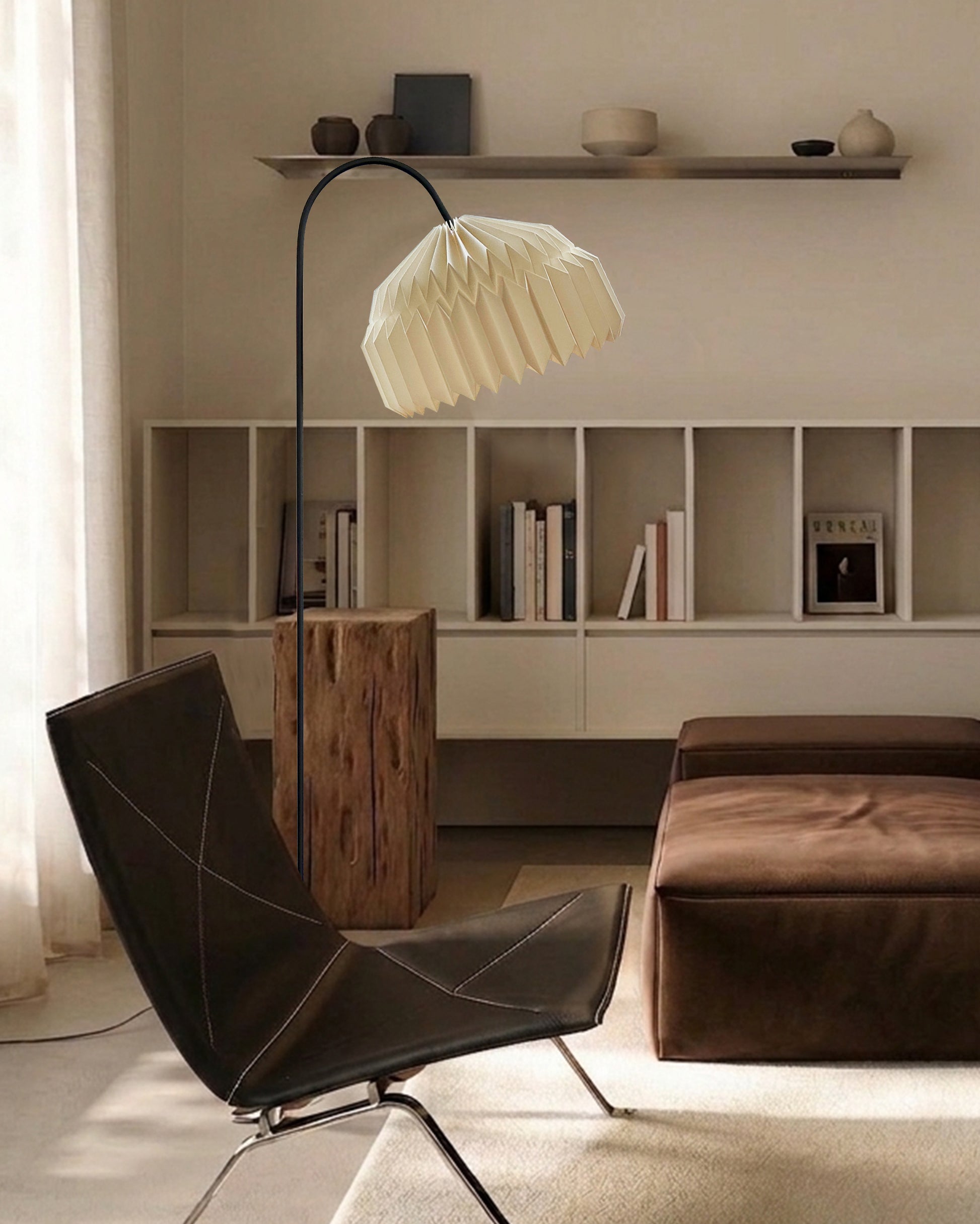Plié Lights, lámpara de piso en forma de arco hecha en colombia. La base metálica tiene un tamaño perfecto para lámpara de lectura. La caperuza es referencia Alfa, tiene un diseño que genera luz directa hacia abajo. El diseño tiene pliegues en papel geométricos. Produce una iluminación cálida.Fotografía de producto tomada en sitio en una sala, en primer plano se ve una silla y al lado derecho un puff en cuero. En el fondo una biblioteca con libros. La escena es de día y la lámpara está apagada. 