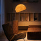 Plié Lights, lámpara de piso en forma de arco hecha en colombia. La base metálica tiene un tamaño perfecto para lámpara de lectura. La caperuza es referencia Alfa, tiene un diseño que genera luz directa hacia abajo. El diseño tiene pliegues en papel geométricos. Produce una iluminación cálida.Fotografía de producto tomada en sitio en una sala, en primer plano se ve una silla y al lado derecho un puff en cuero. En el fondo una biblioteca con libros. La escena es nocturna y la lámpara está prendida.