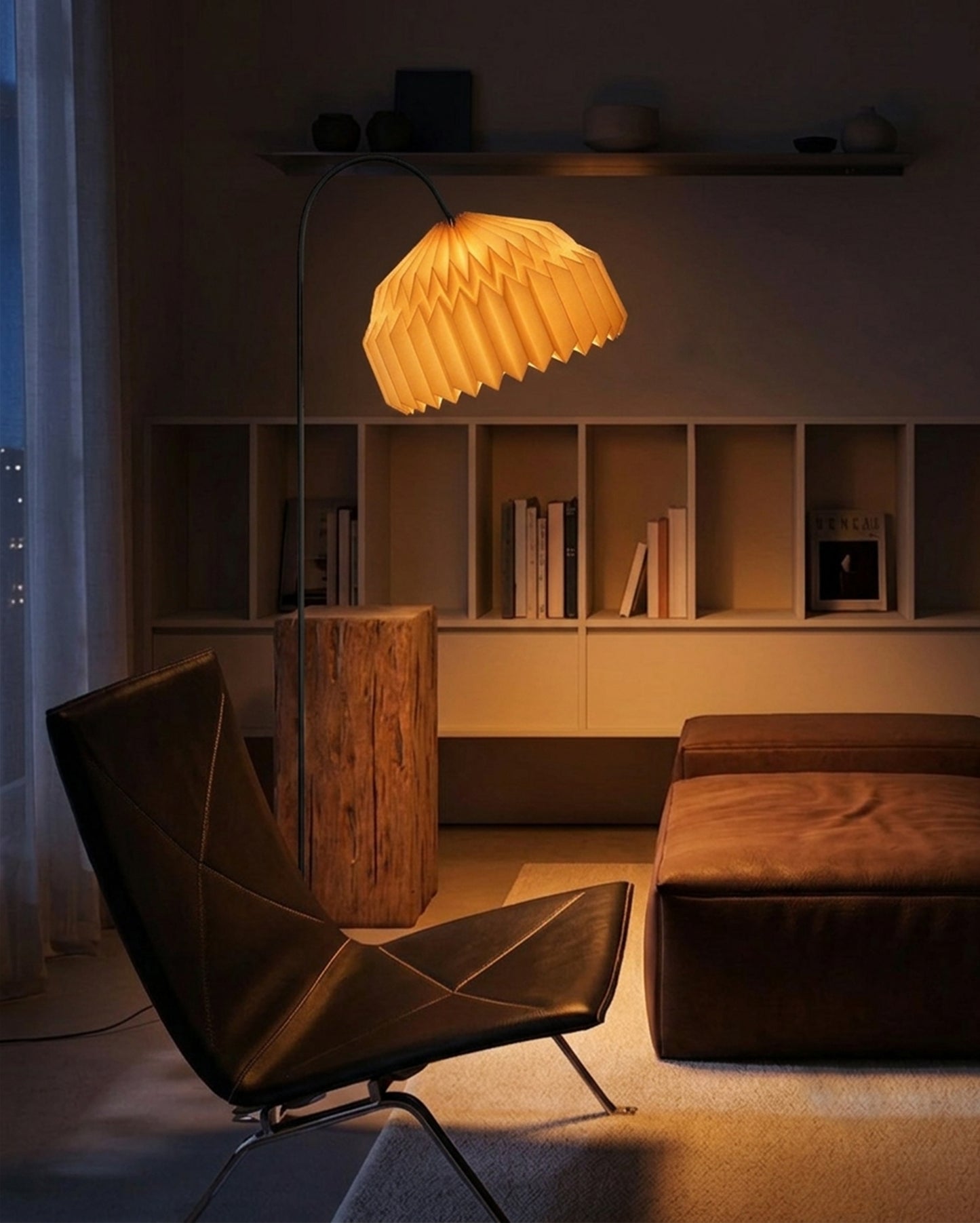 Plié Lights, lámpara de piso en forma de arco hecha en colombia. La base metálica tiene un tamaño perfecto para lámpara de lectura. La caperuza es referencia Alfa, tiene un diseño que genera luz directa hacia abajo. El diseño tiene pliegues en papel geométricos. Produce una iluminación cálida.Fotografía de producto tomada en sitio en una sala, en primer plano se ve una silla y al lado derecho un puff en cuero. En el fondo una biblioteca con libros. La escena es nocturna y la lámpara está prendida.