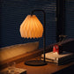 Plié Lights_Lámparas de mesa hechas en Colombia_Caperuzas con diseños geométricos hechas con papel. Fotografía de producto puesta encima de una consola, a los lados unos libros, un baso y un florero. La escena es nocturna y la lámpara está prendida generando un ambiente cálido. 