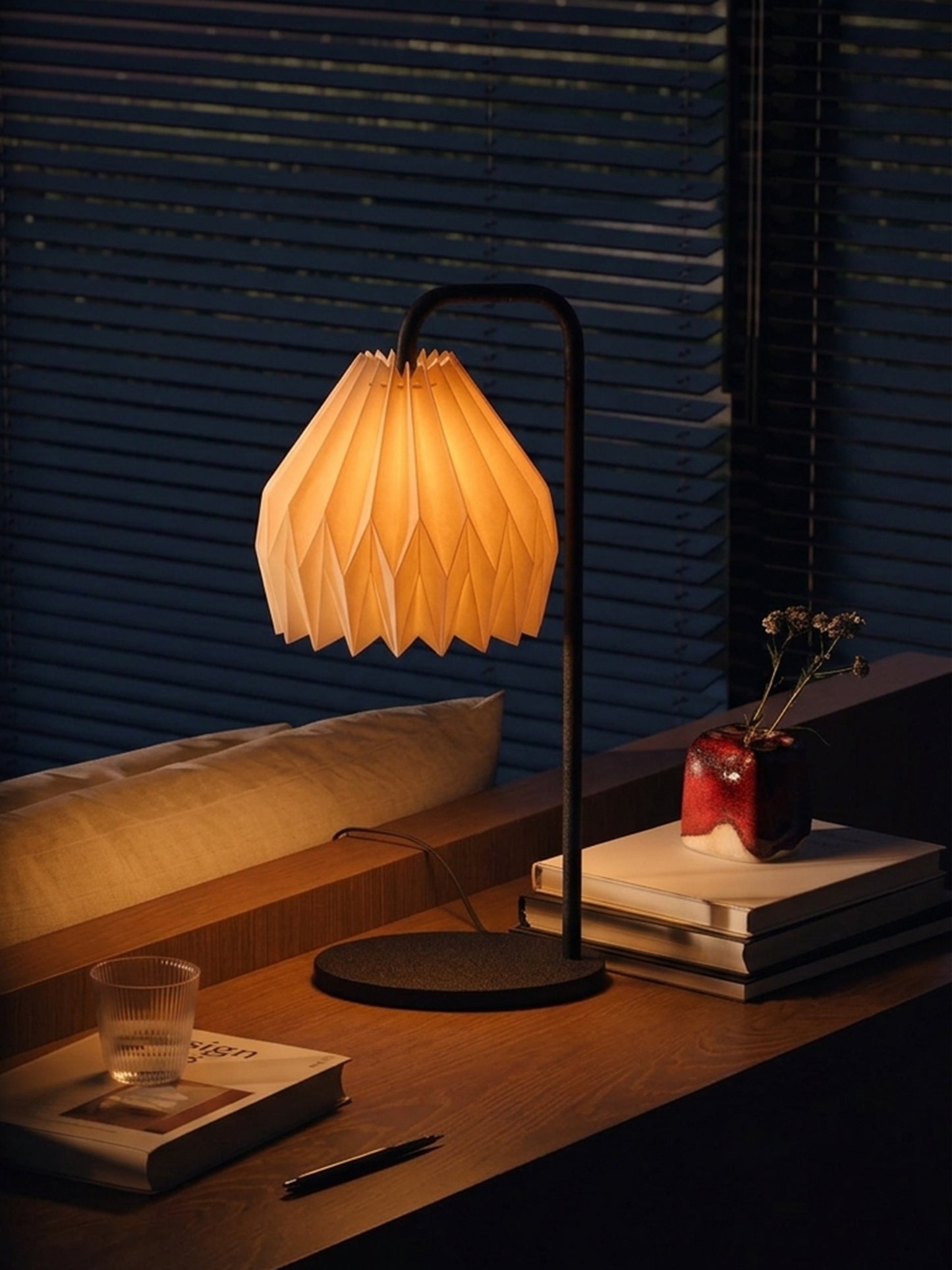 Plié Lights_Lámparas de mesa hechas en Colombia_Caperuzas con diseños geométricos hechas con papel. Fotografía de producto puesta encima de una consola, a los lados unos libros, un baso y un florero. La escena es nocturna y la lámpara está prendida generando un ambiente cálido. 