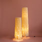 Lámparas de piso de Colombia. Plié Lights, referencia  Torre L y XL de la colección monumentos. Fotografía de producto con fondo de color rosa. Caperuza inspirada en arquitectura newyorkina. El papel tiene cortes lo que permite genera movimiento entre luz directa e indirecta. Hecha en papel con mano 100% colombianas. La base metálica es color negro y cable negro. Las lámparas están prendidas. 