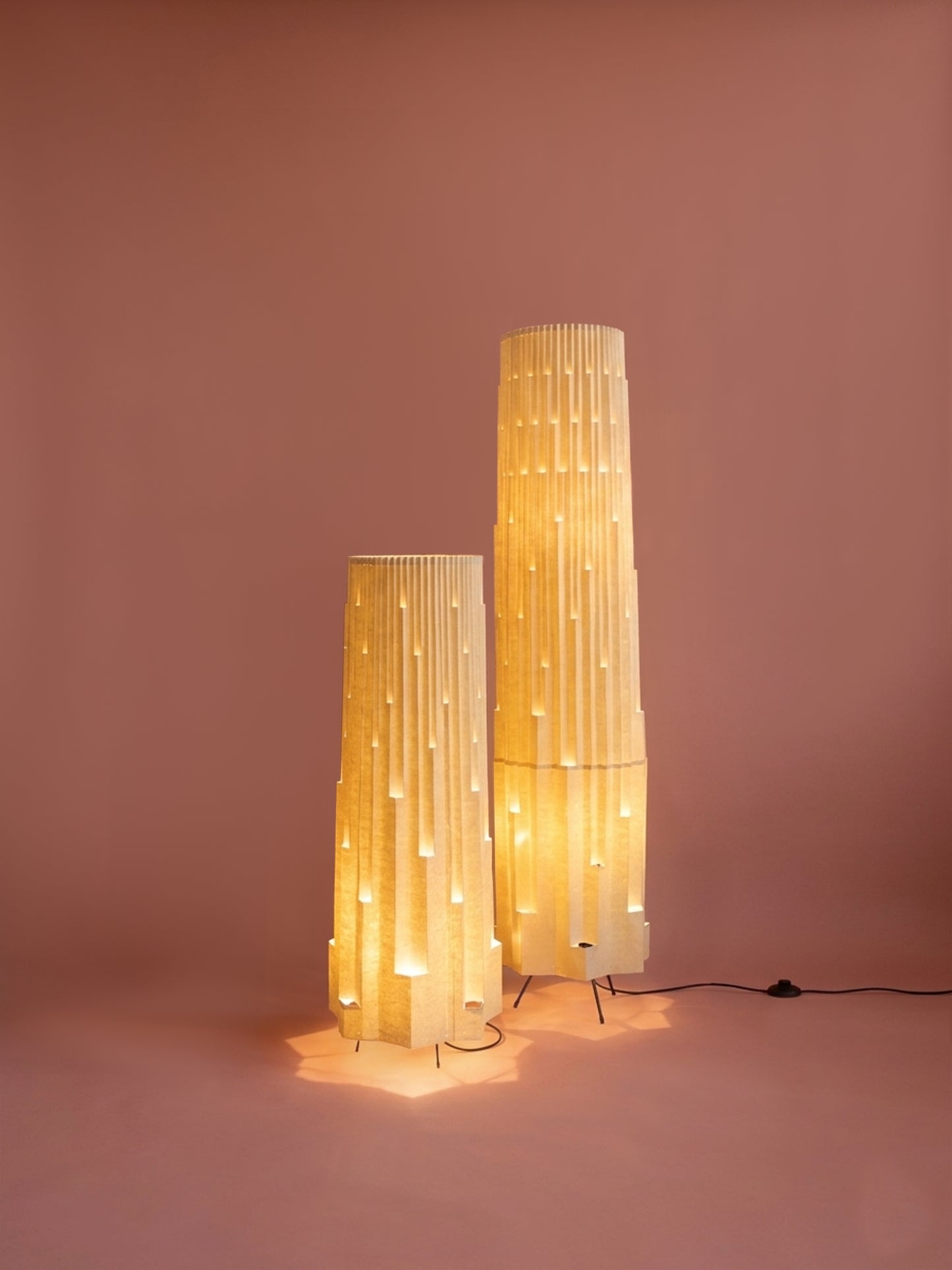 Lámparas de piso de Colombia. Plié Lights, referencia  Torre L y XL de la colección monumentos. Fotografía de producto con fondo de color rosa. Caperuza inspirada en arquitectura newyorkina. El papel tiene cortes lo que permite genera movimiento entre luz directa e indirecta. Hecha en papel con mano 100% colombianas. La base metálica es color negro y cable negro. Las lámparas están prendidas. 
