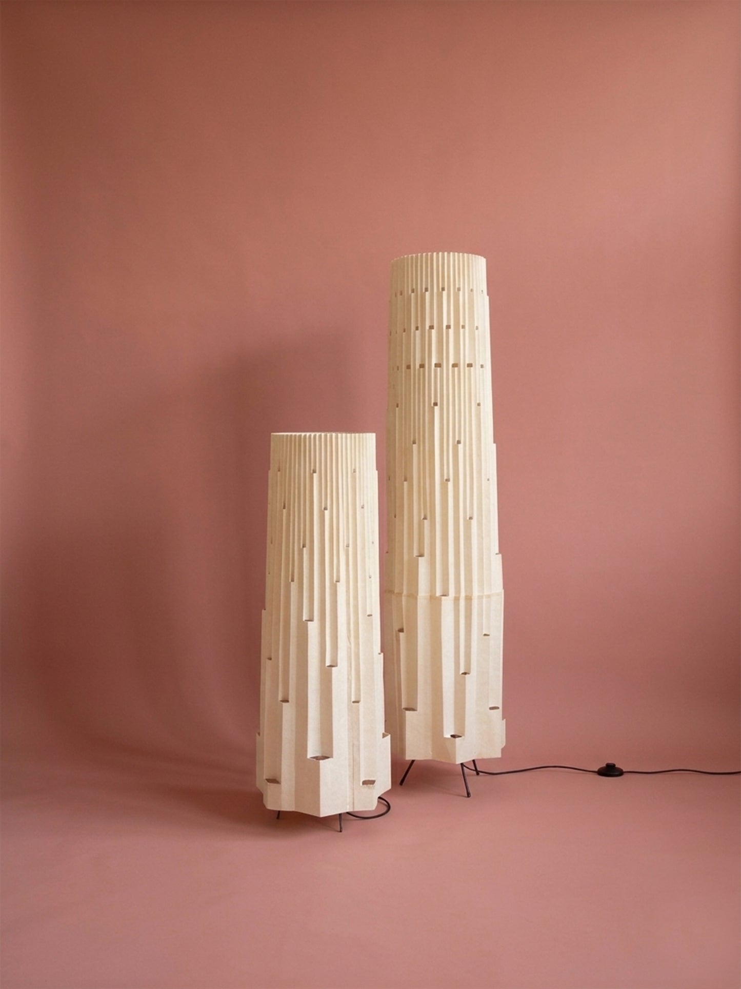 Lámparas de piso de Colombia. Plié Lights, referencia  Torre L y XL de la colección monumentos. Fotografía de producto con fondo de color rosa. Caperuza inspirada en arquitectura newyorkina. El papel tiene cortes lo que permite genera movimiento entre luz directa e indirecta. Hecha en papel con mano 100% colombianas. La base metálica es color negro y cable negro. Las lámparas están apagadas. 