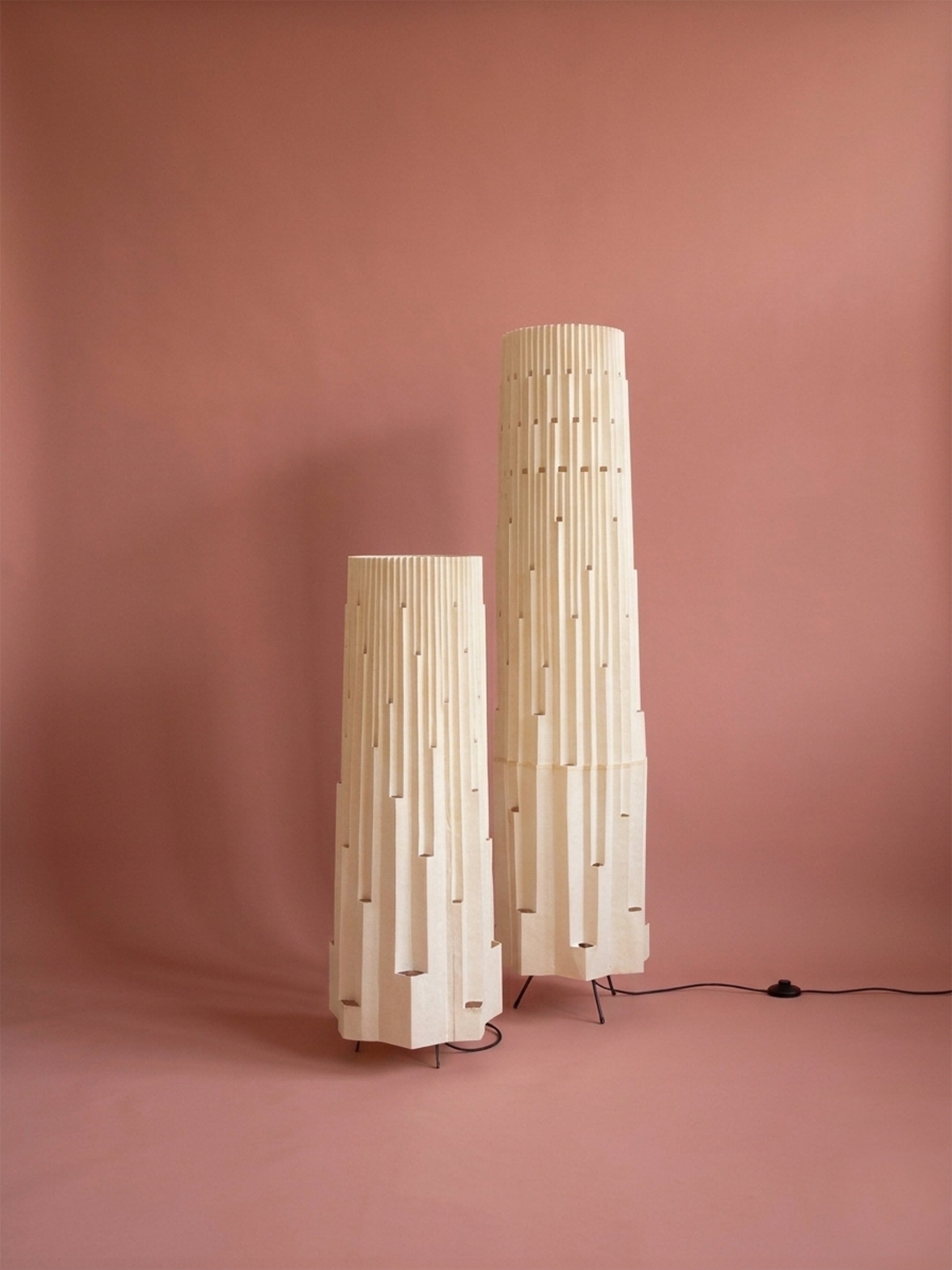 Lámparas de piso de Colombia. Plié Lights, referencia  Torre L y XL de la colección monumentos. Fotografía de producto con fondo de color rosa. Caperuza inspirada en arquitectura newyorkina. El papel tiene cortes lo que permite genera movimiento entre luz directa e indirecta. Hecha en papel con mano 100% colombianas. La base metálica es color negro y cable negro. Las lámparas están apagadas. 
