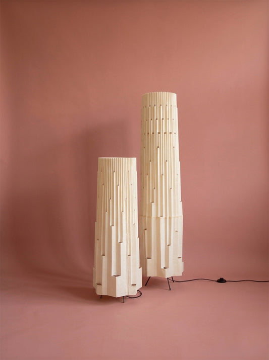 Lámparas de piso de Colombia. Plié Lights, referencia  Torre L y XL de la colección monumentos. Fotografía de producto con fondo de color rosa. Caperuza inspirada en arquitectura newyorkina. El papel tiene cortes lo que permite genera movimiento entre luz directa e indirecta. Hecha en papel con mano 100% colombianas. La base metálica es color negro y cable negro. Las lámparas están apagadas. 