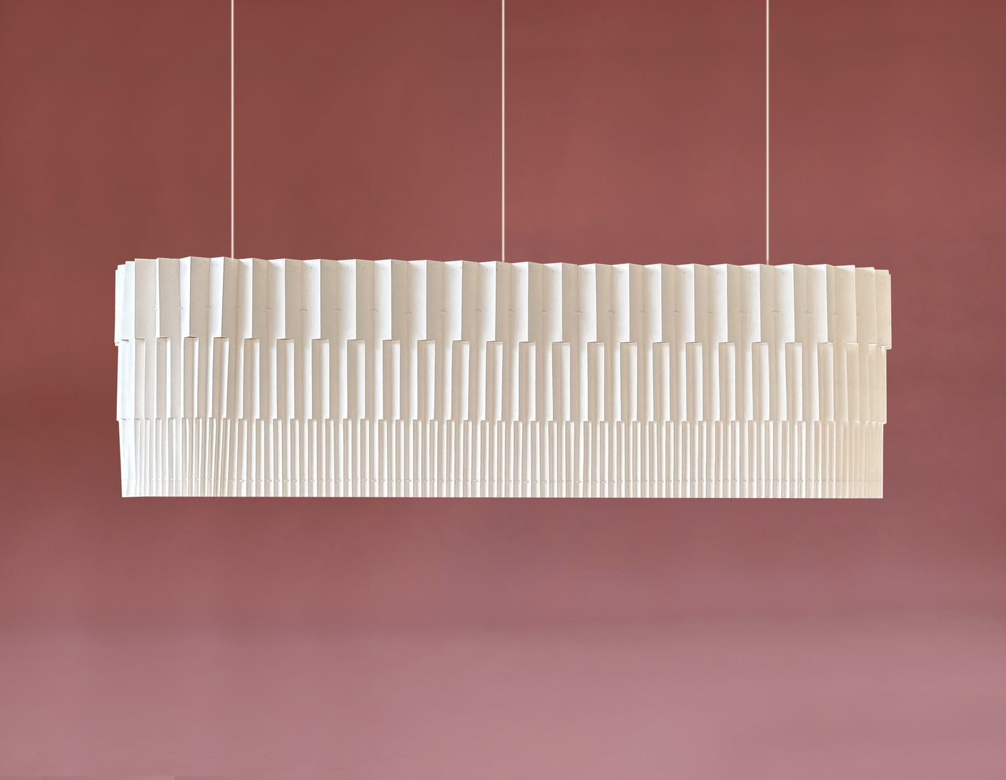 Lámpara colgante de Colombia. Plié Lights, referencia Capitolio de la colección monumentos. Fotografía de producto tomada sobre un fondo color ropa. La lámpara es ancha ó larga de forma rectanculas de bordes redondeados. El diseño del papel plegado también tiene cortes que permiten que la luz pasé directamente através y se genere una iluminación como la de un edificio arquitectónico. El papel tiene cortes lo que permite genera movimiento entre luz directa e indirecta. Hecha en papel a mano.