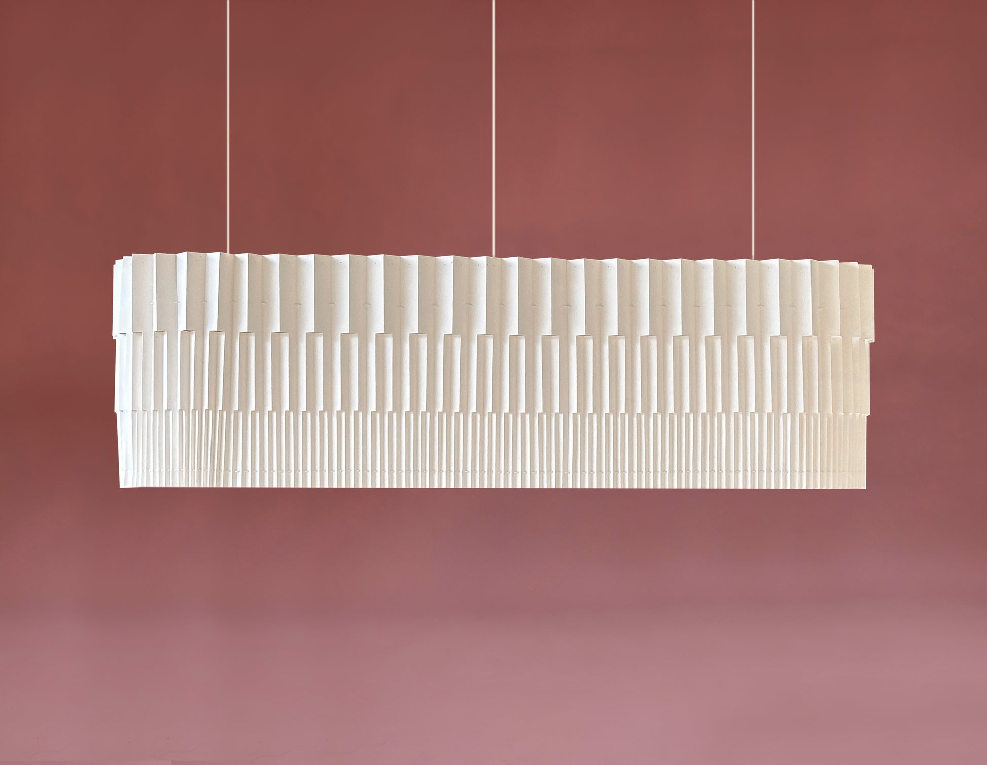 Lámpara colgante de Colombia. Plié Lights, referencia Capitolio de la colección monumentos. Fotografía de producto tomada sobre un fondo color ropa. La lámpara es ancha ó larga de forma rectanculas de bordes redondeados. El diseño del papel plegado también tiene cortes que permiten que la luz pasé directamente através y se genere una iluminación como la de un edificio arquitectónico. El papel tiene cortes lo que permite genera movimiento entre luz directa e indirecta. Hecha en papel a mano.