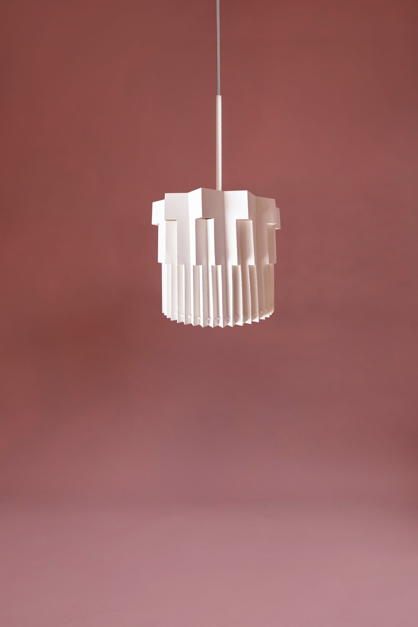 Lámpara colgante de Colombia. Plié Lights, referencia  Fortaleza de la colección Monumentos. Fotografía de producto sobre un fondo color rosa base metálica color blanca. Caperuza de papel doblada con cortes que generan una iluminación arquitectónica nocturna. Hecha en papel a mano. La base metálica es color negro y cable negro.