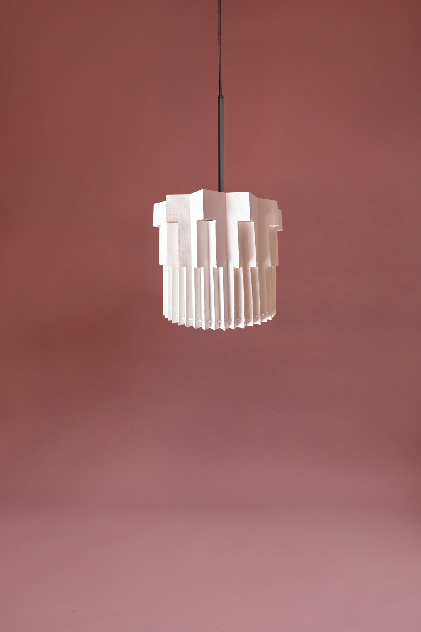 Lámpara colgante de Colombia. Plié Lights, referencia  Fortaleza de la colección Monumentos. Fotografía de producto sobre un fondo color rosa base metálica color negra. Caperuza de papel doblada con cortes que generan una iluminación arquitectónica nocturna. Hecha en papel a mano. La base metálica es color negro y cable negro.