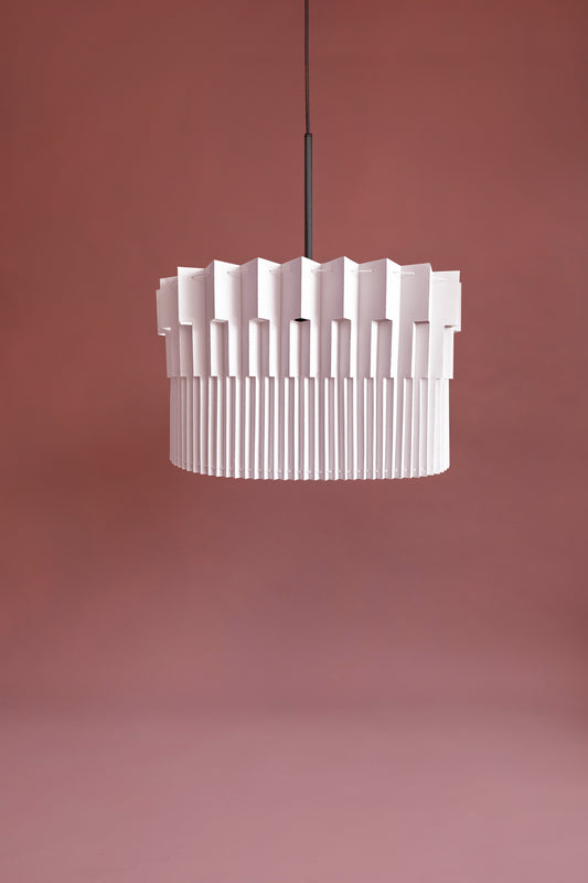 Lámpara colgante de Colombia. Plié Lights, referencia Museo de la colección monumentos. Fotografía de producto con fondo de color rosa. Caperuza escultórica estilo arquitectónico. El papel tiene cortes lo que permite genera movimiento entre luz directa e indirecta. Hecha en papel a mano. La base metálica es color negro y cable negro.