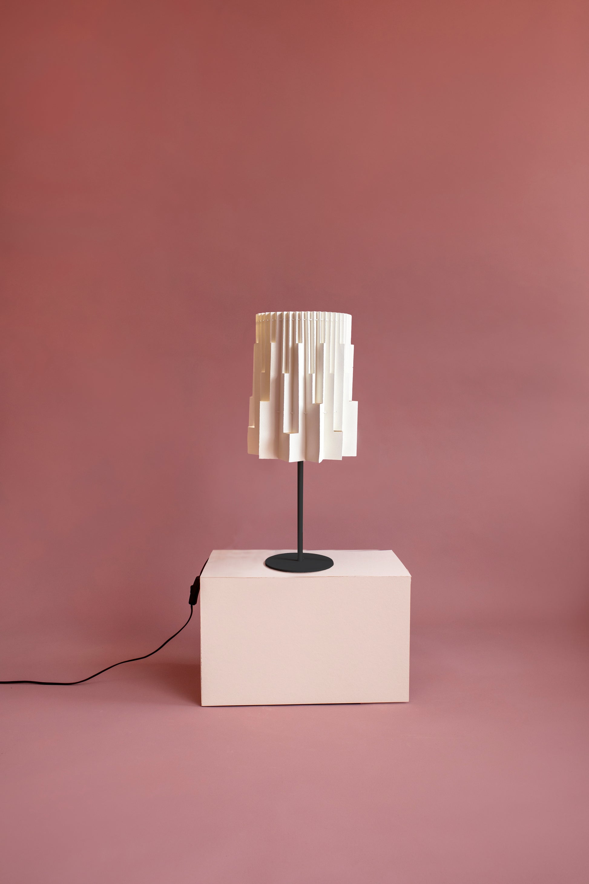 Plié Lights, lámpara de mesa referencia Pilar, de la colección Monumentos, hecha a mano, caperuza hecha en papel plegado a mano. Soporte metálica y cable negro Acabado negro. Producto hecho en Colombia. Diseño Colombiano. Figura geométrica muy interesante. Ideal para iluminar y darle calidez a los espacios. efecto de luz y sombras generado por cortes en el papel que dejan pasar luz directa.